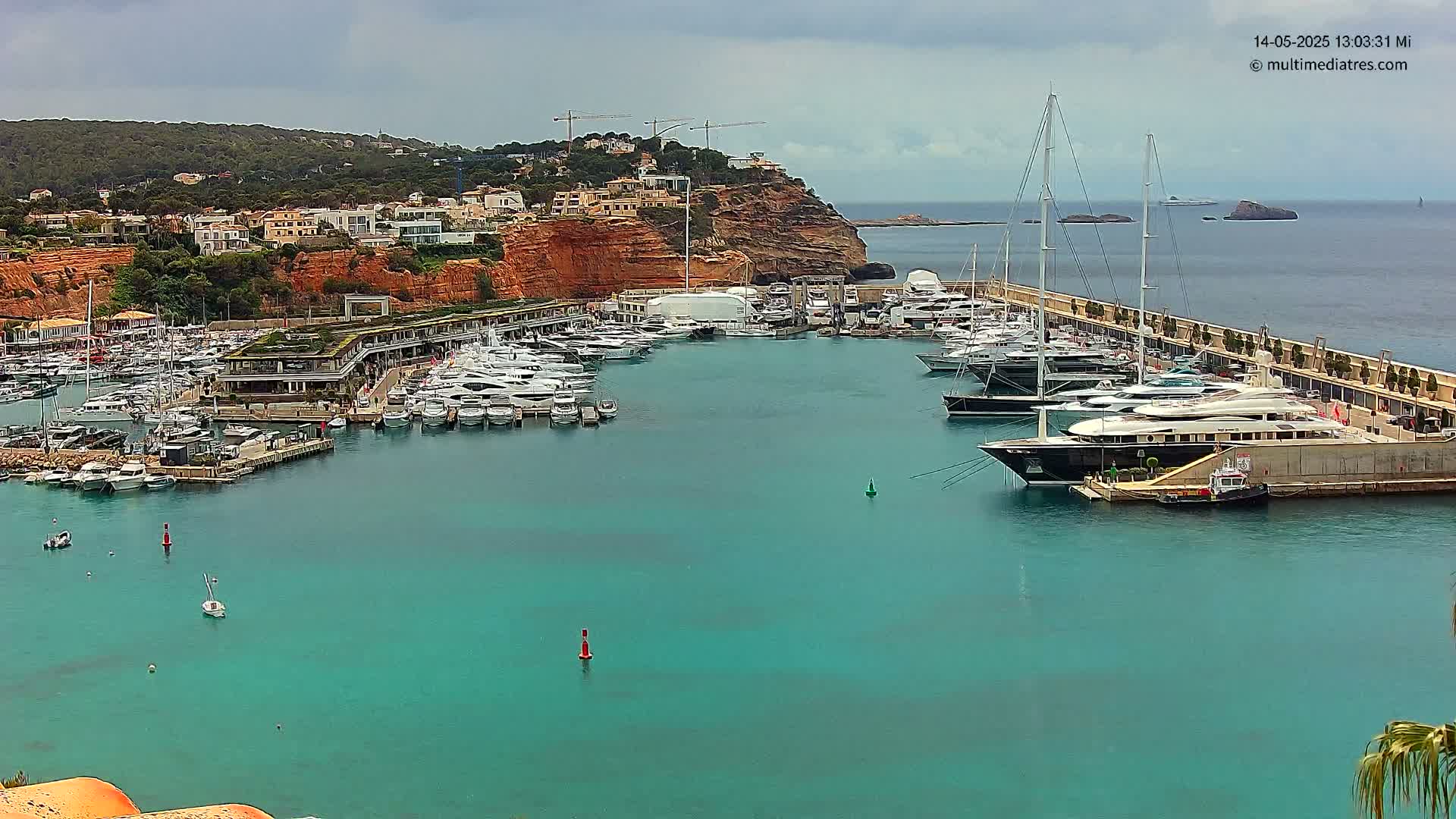 A marina filled with yachts and boats is nestled against a red cliffside overlooking a turquoise sea under a cloudy sky.
