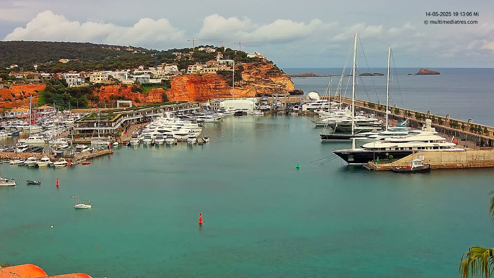 A marina filled with yachts and boats is nestled beside a red-cliff hillside under a partly cloudy sky.