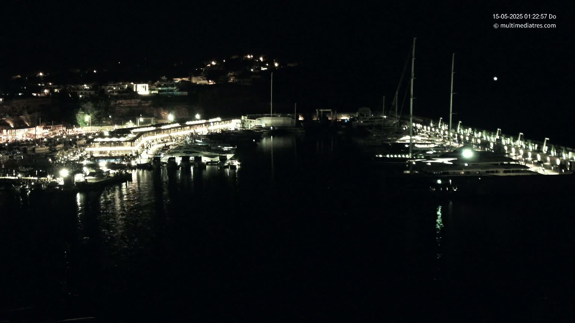 A nighttime harbor scene shows numerous boats docked along a waterfront lined with illuminated buildings, under a clear, dark sky.