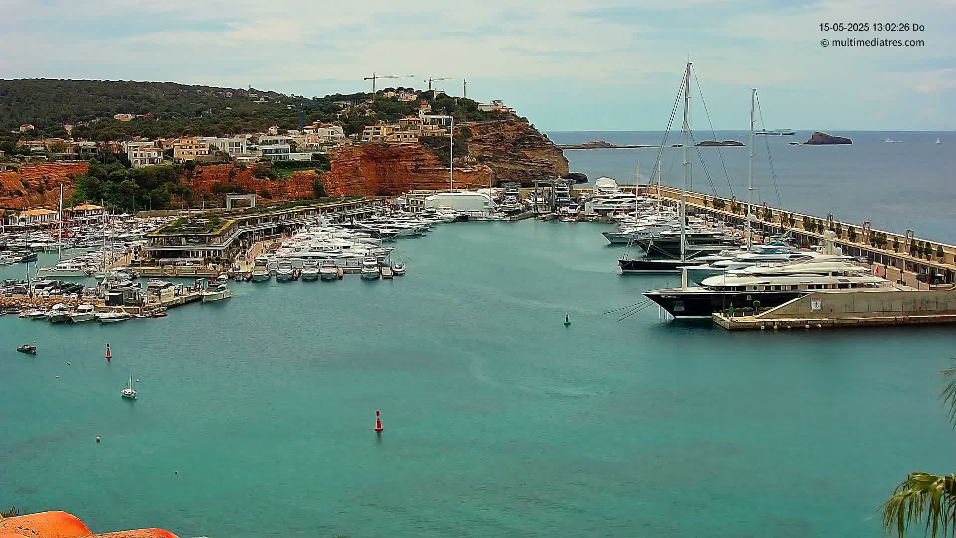 A marina filled with numerous yachts and boats is nestled against a red-cliffed hillside under a partly cloudy sky.