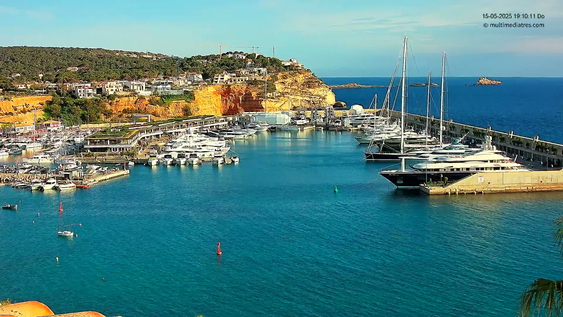 A marina filled with yachts and boats is nestled against a hillside of buildings under a clear blue sky.