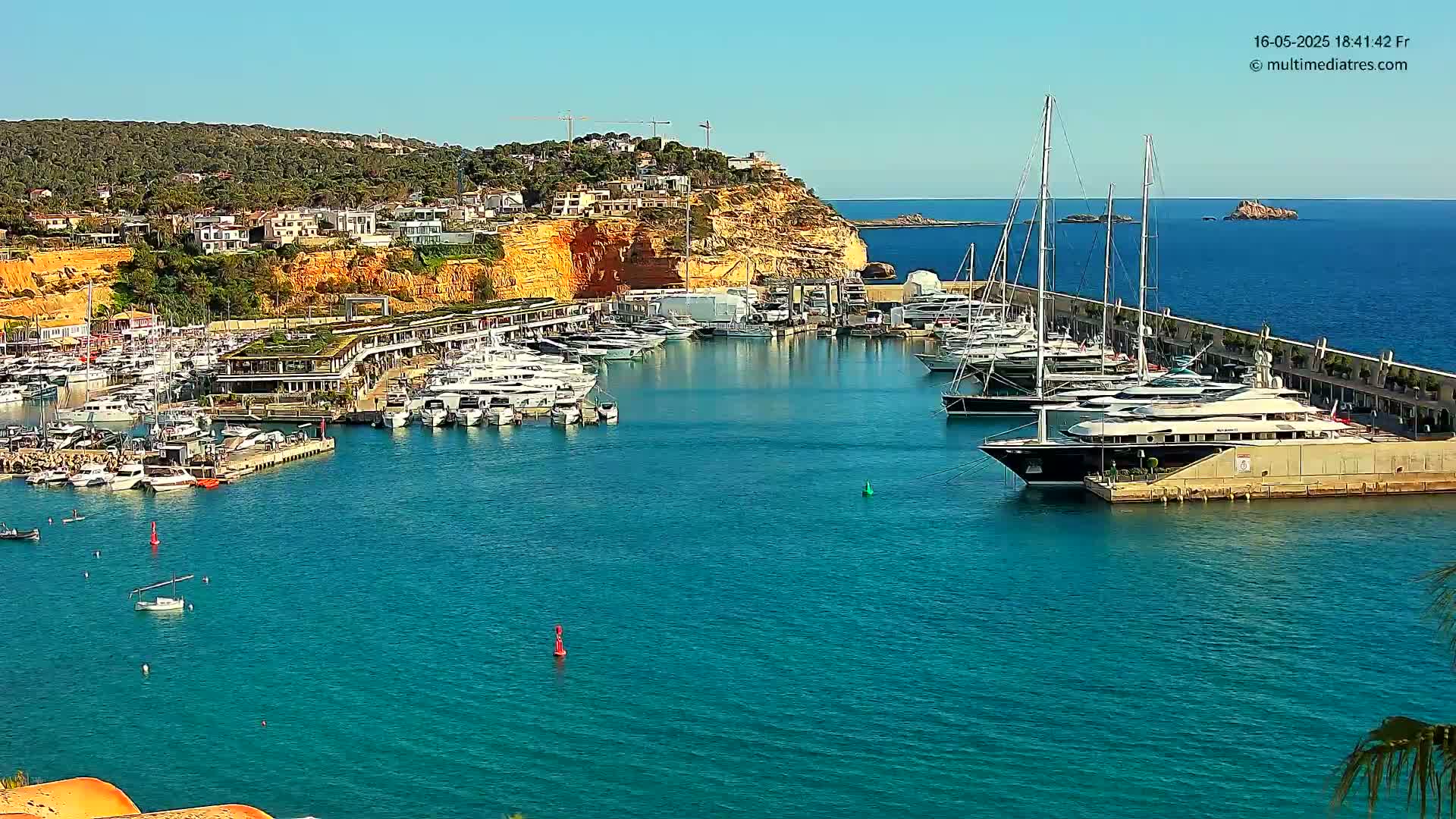 A marina filled with numerous yachts and boats is situated along a clear blue body of water next to a hillside of buildings under a sunny sky.