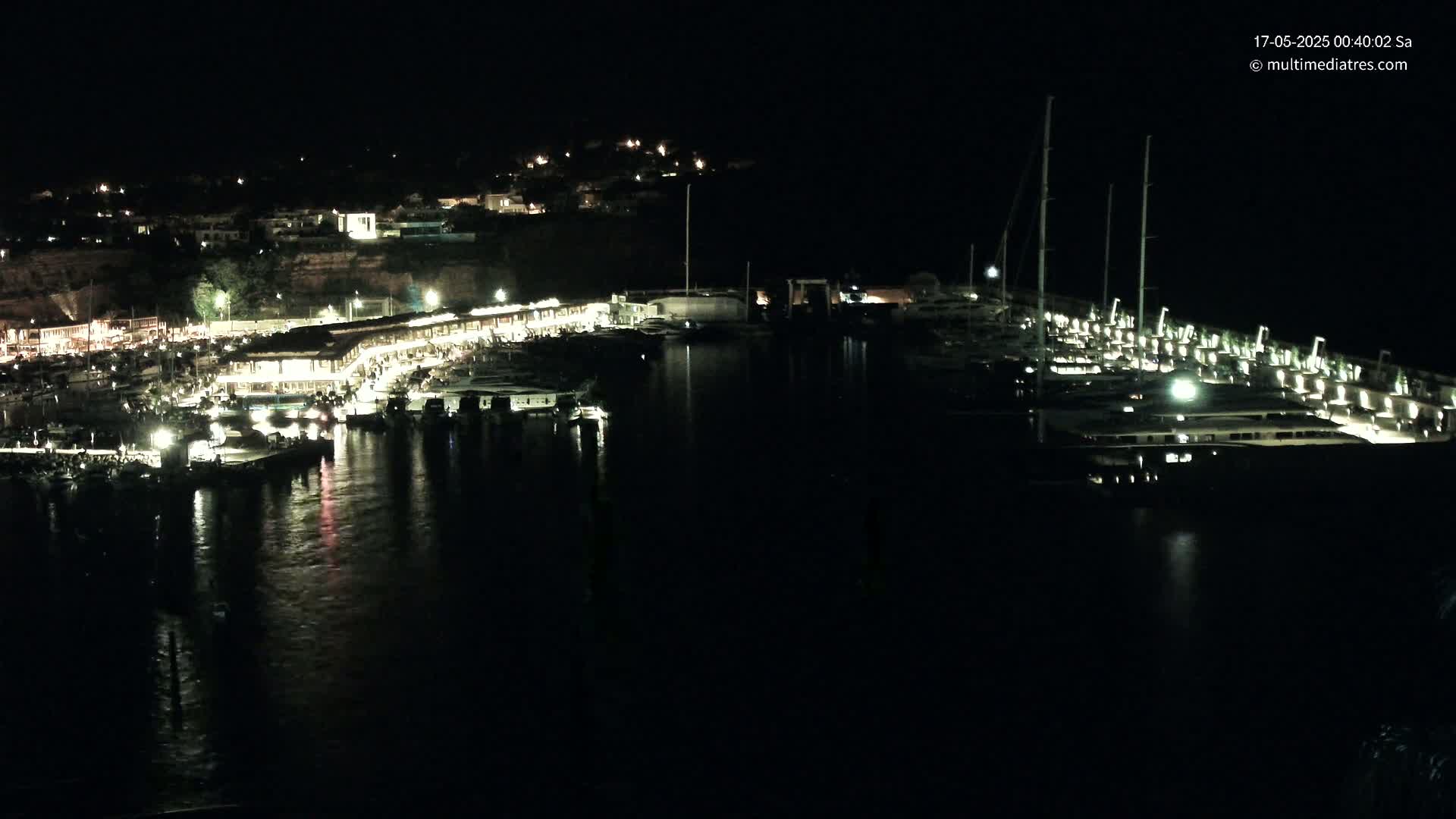 A nighttime view of a marina filled with boats, with lights illuminating the docks and surrounding buildings on a calm body of water.