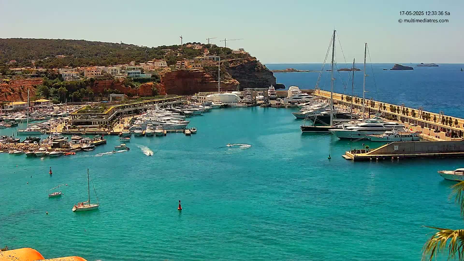 A marina filled with yachts and sailboats is nestled into a coastal town on a sunny day, with clear blue water stretching to distant islands.
