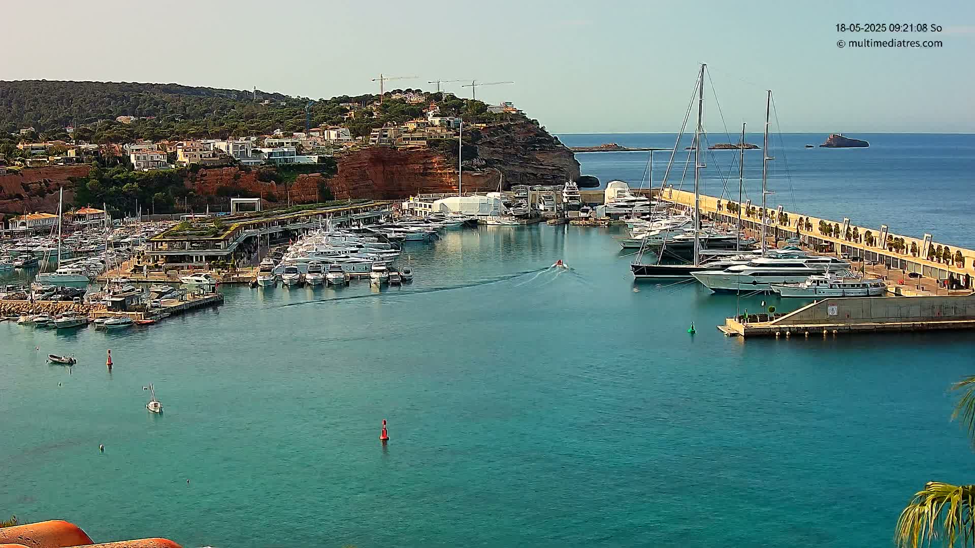 A marina filled with numerous yachts and boats is nestled in a calm, clear blue harbor, with a hillside of buildings visible in the background under a sunny sky.