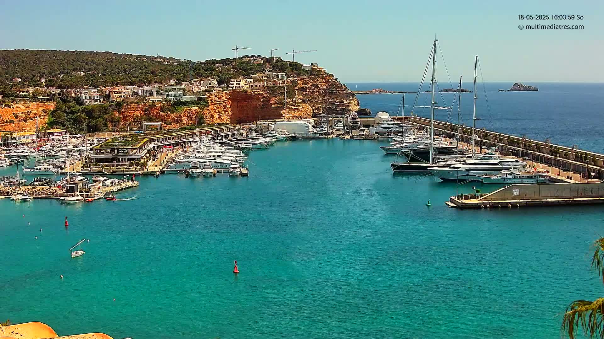 A marina filled with yachts and boats is nestled into a hillside next to a clear, turquoise body of water under a sunny sky.