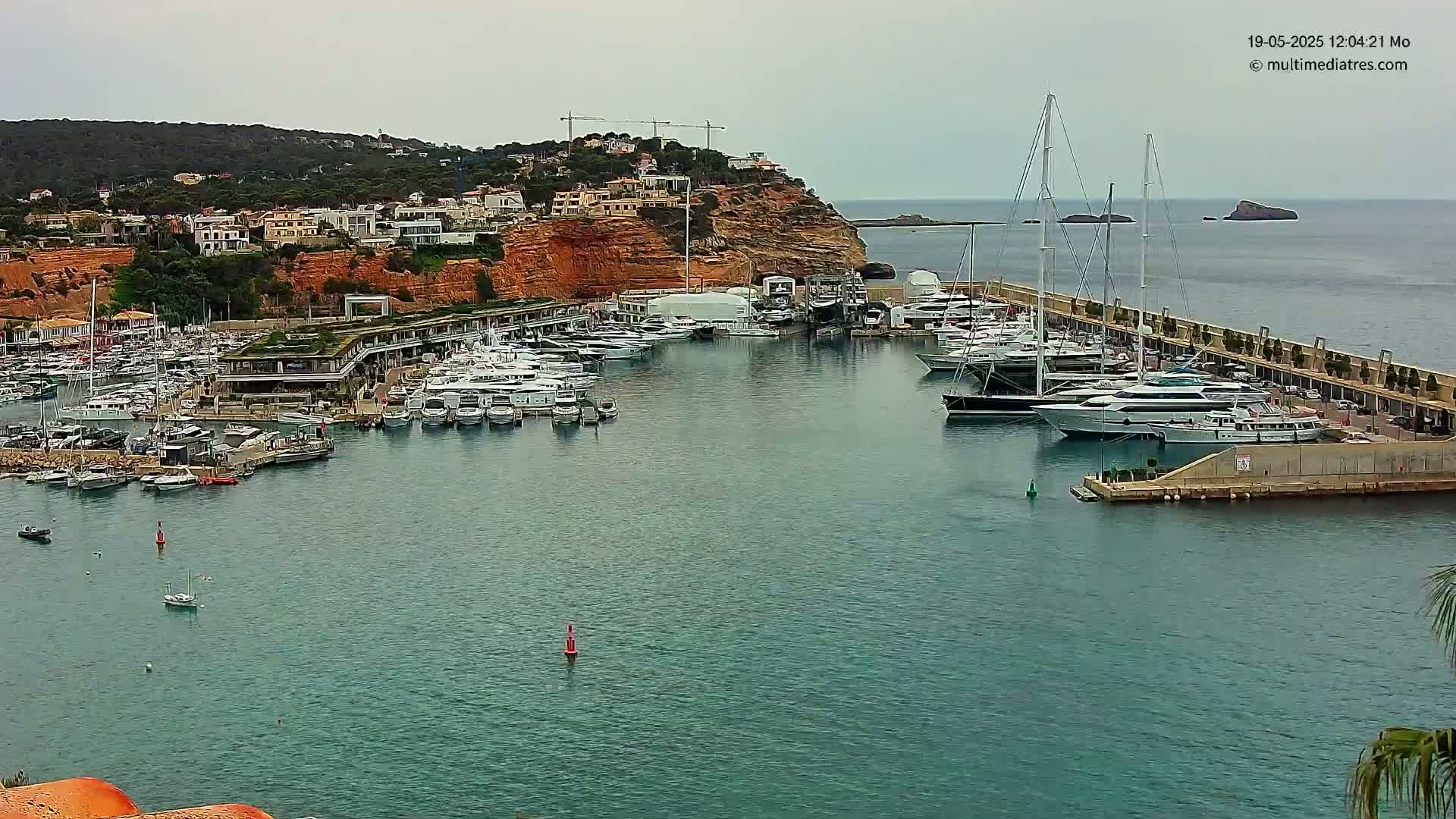 A marina filled with numerous yachts and boats is nestled against a rocky, reddish-brown hillside under an overcast sky.