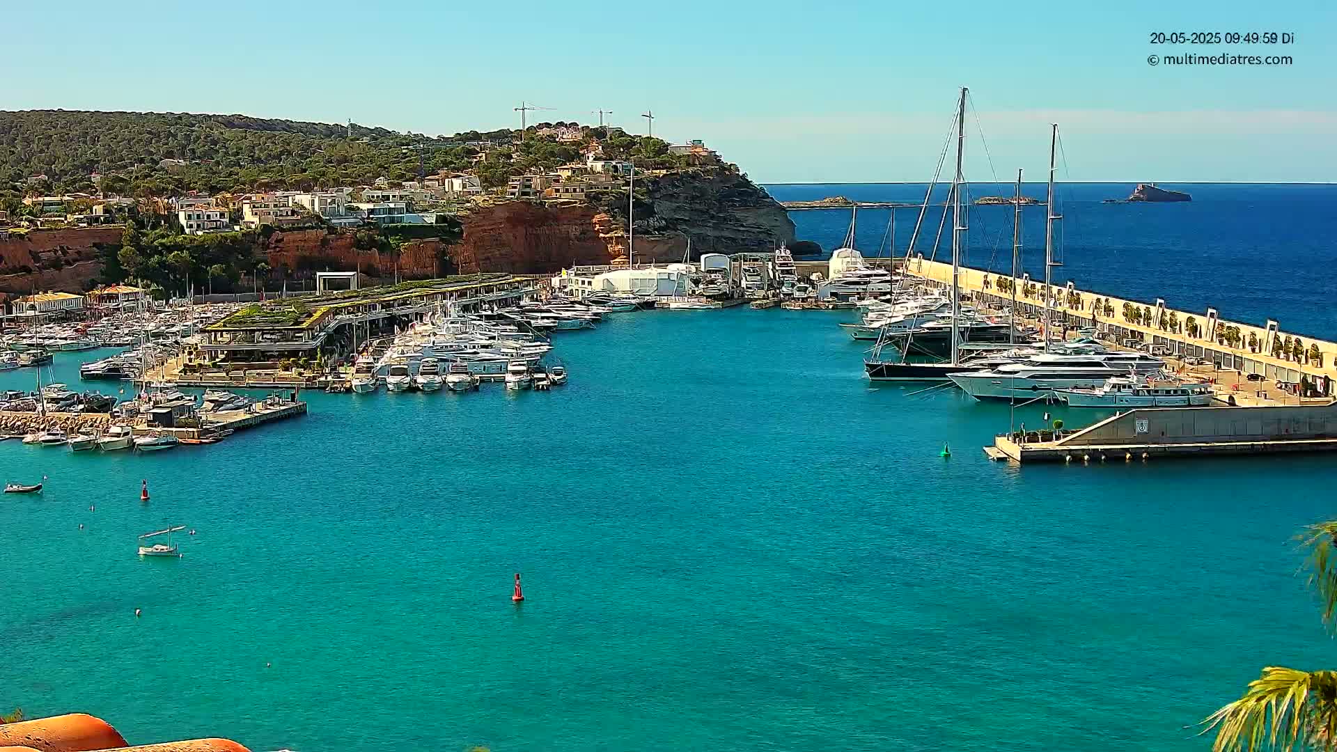 A marina filled with numerous yachts and boats is situated in a harbor next to a hillside town under a clear, sunny sky.