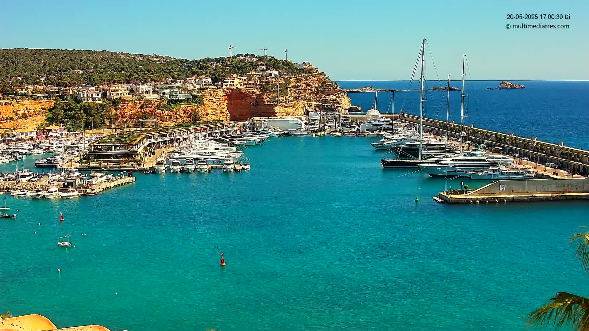 A marina filled with yachts and boats is situated on a clear, sunny day next to a rocky, Mediterranean-style coastline with residential buildings.