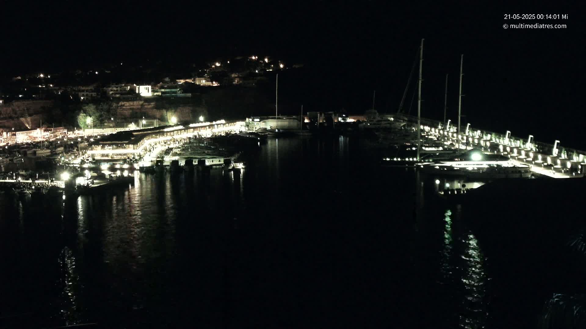 A nighttime view of a marina filled with boats, lit by artificial lights, with a town visible on a hillside in the background.