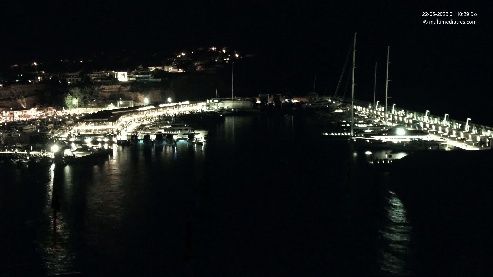 A nighttime view of a harbor filled with boats and yachts, illuminated by lights on the docks and buildings on the shore.