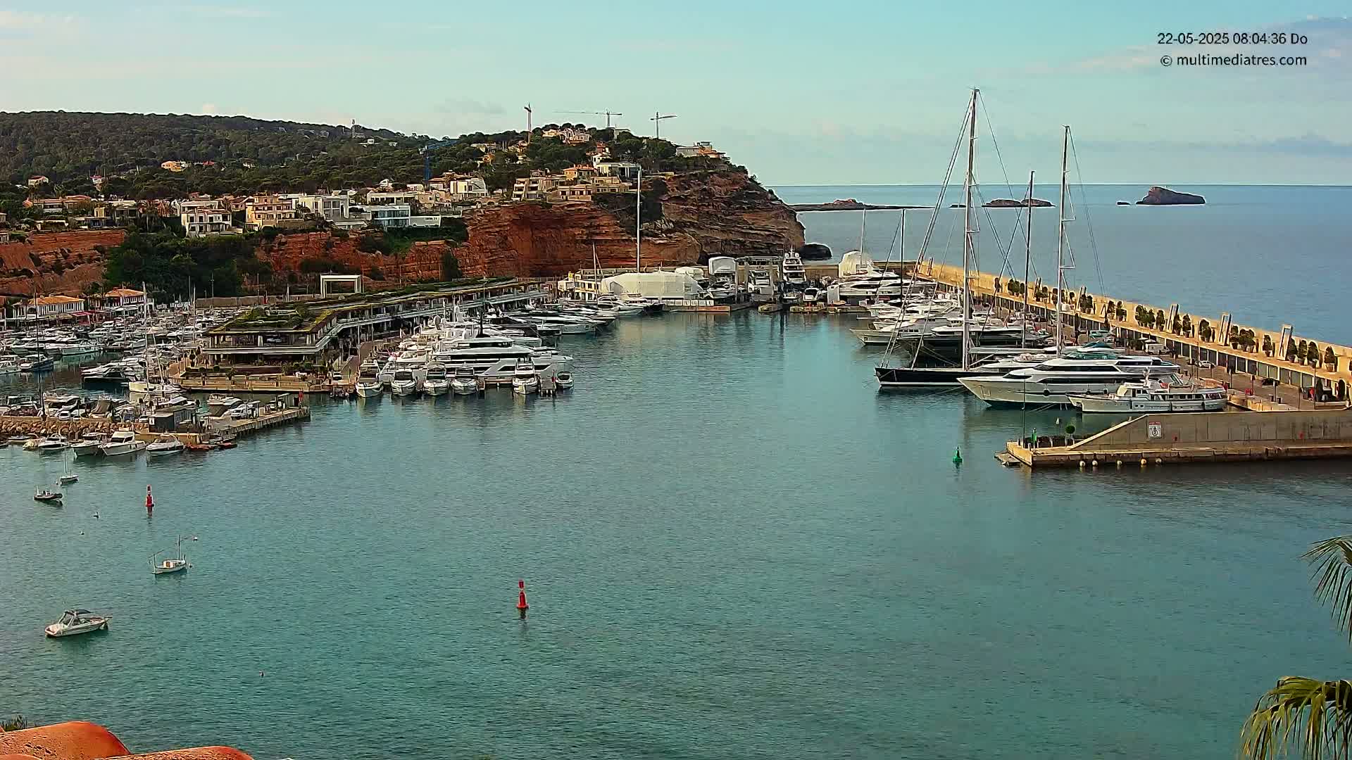A marina filled with yachts and boats is situated along a coastline under a mostly clear sky.