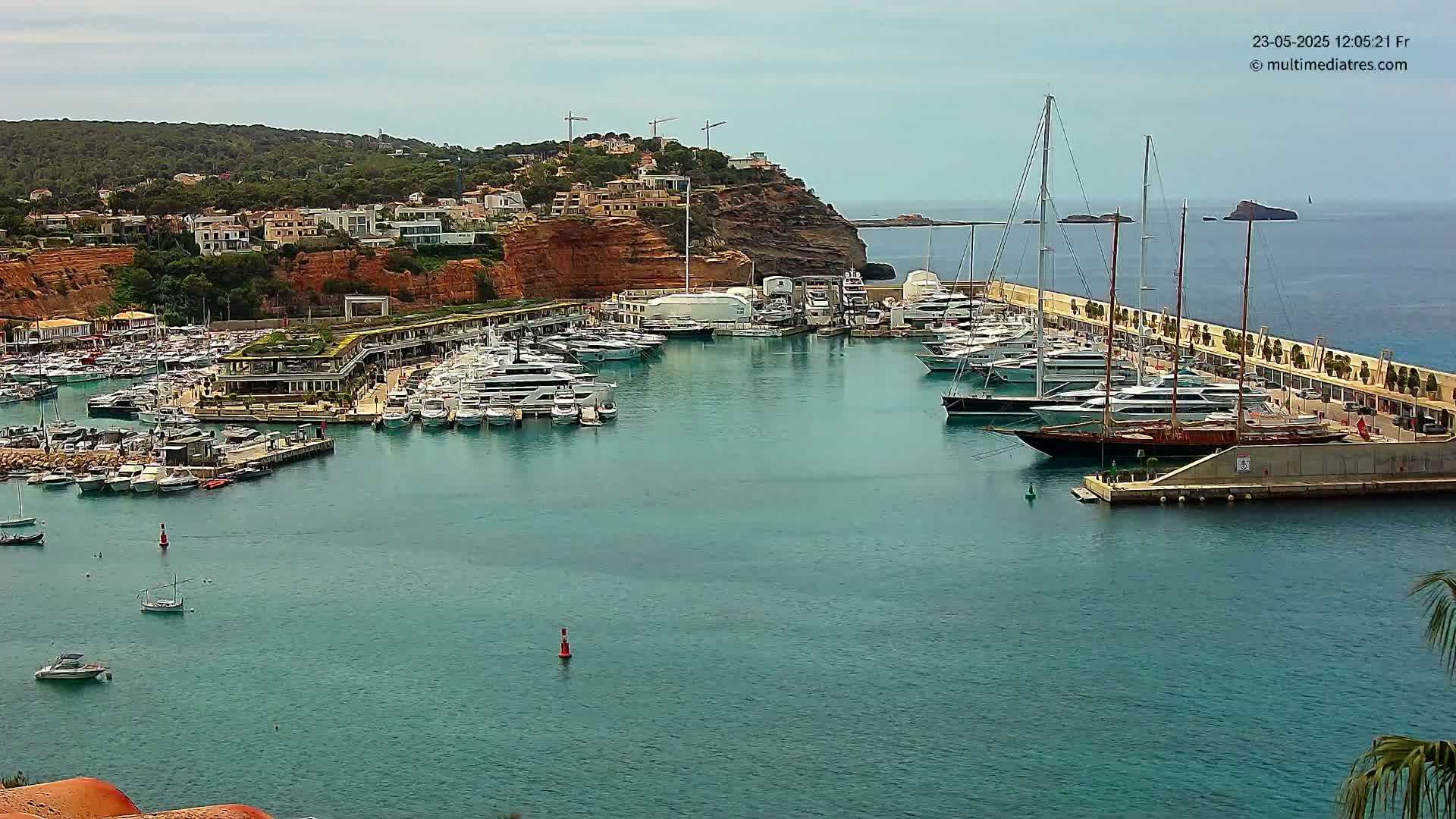 A marina filled with yachts and sailboats is nestled into a hillside overlooking a calm, teal ocean under a mostly clear sky.