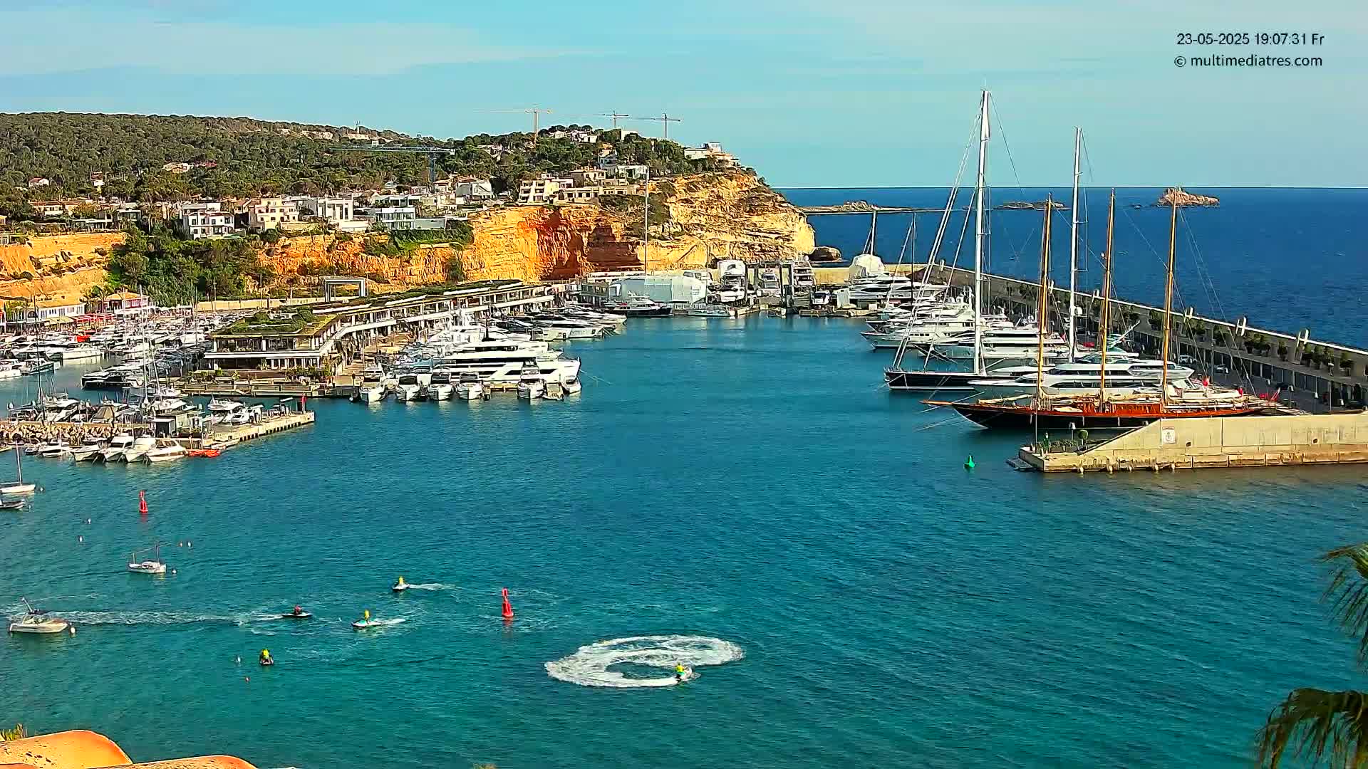 A marina filled with numerous yachts and boats is nestled against a hillside of buildings under a clear, sunny sky.