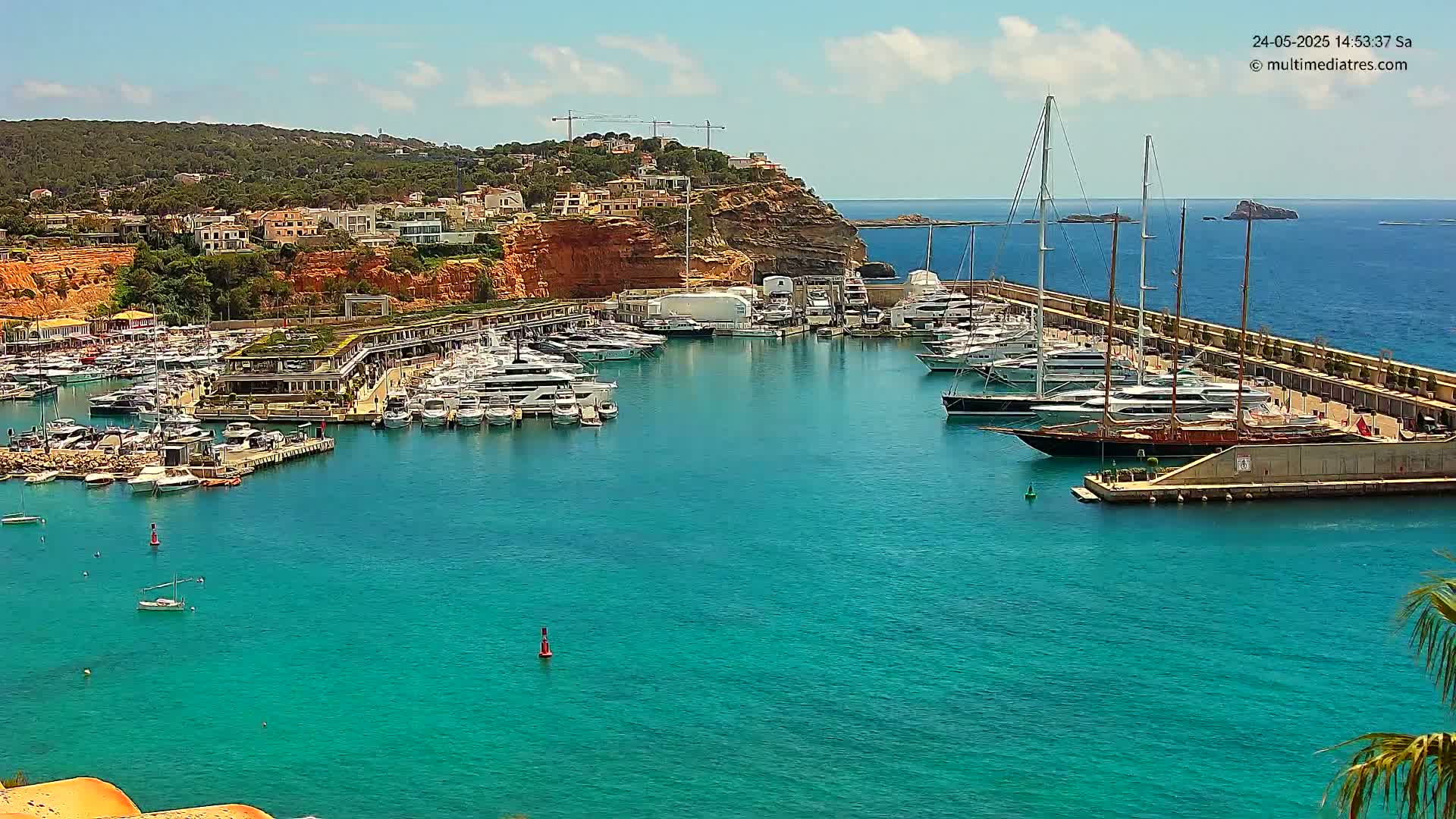 A marina filled with yachts and sailboats is nestled in a calm, turquoise bay, surrounded by a hillside of buildings and reddish cliffs under a mostly sunny sky.