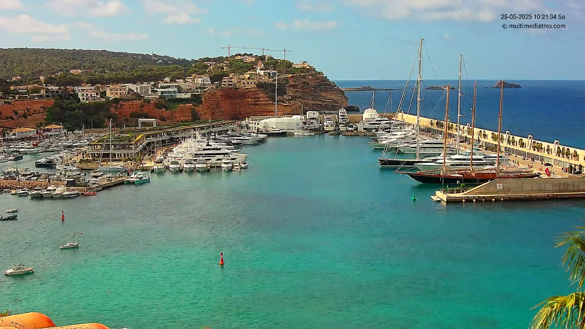 A marina filled with yachts and sailboats is nestled in a turquoise bay, backed by a red cliff topped with houses, under a partly cloudy sky.