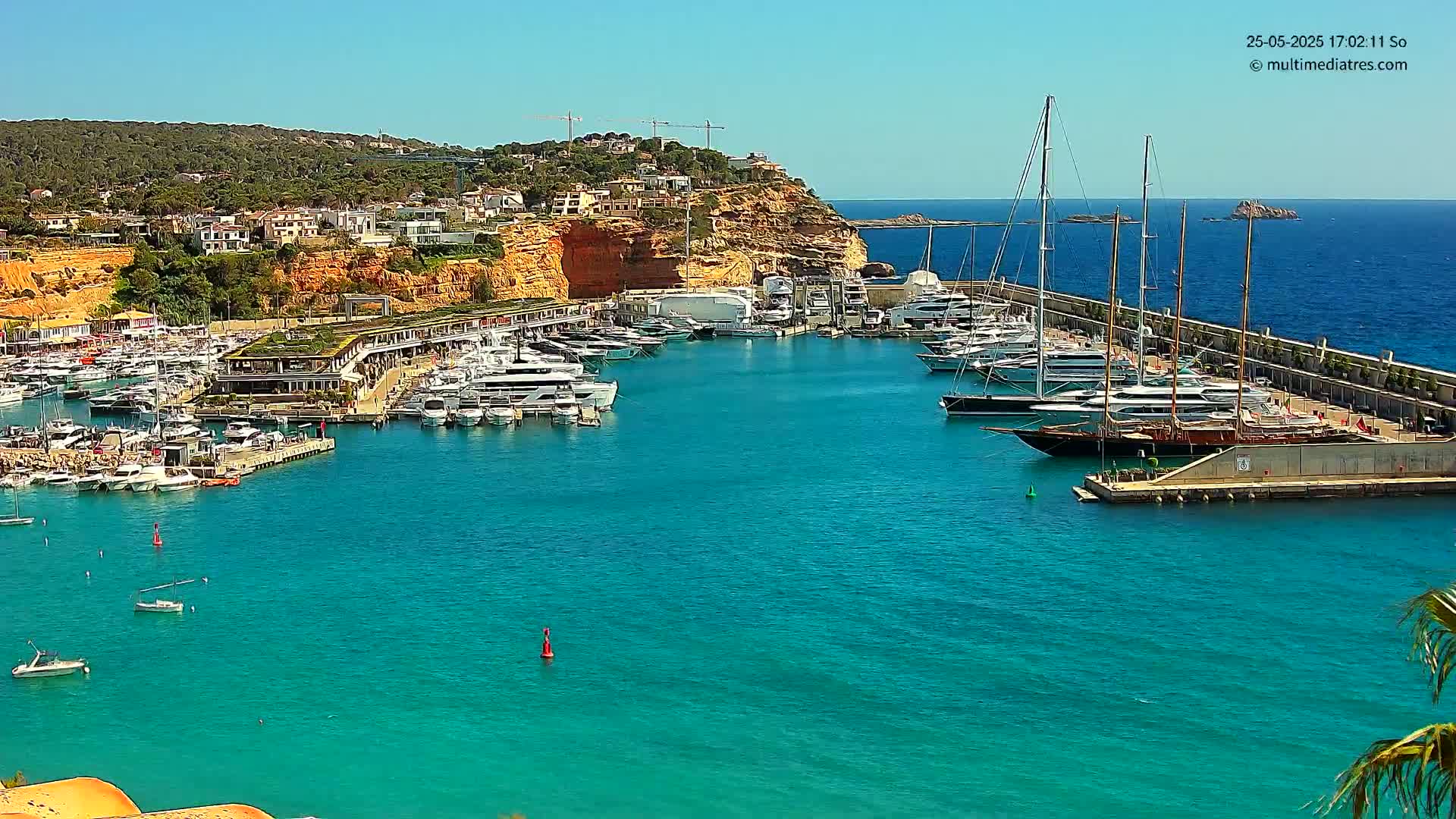 A marina filled with numerous yachts and boats is situated along a clear, turquoise body of water, nestled against a hillside of buildings under a bright, sunny sky.