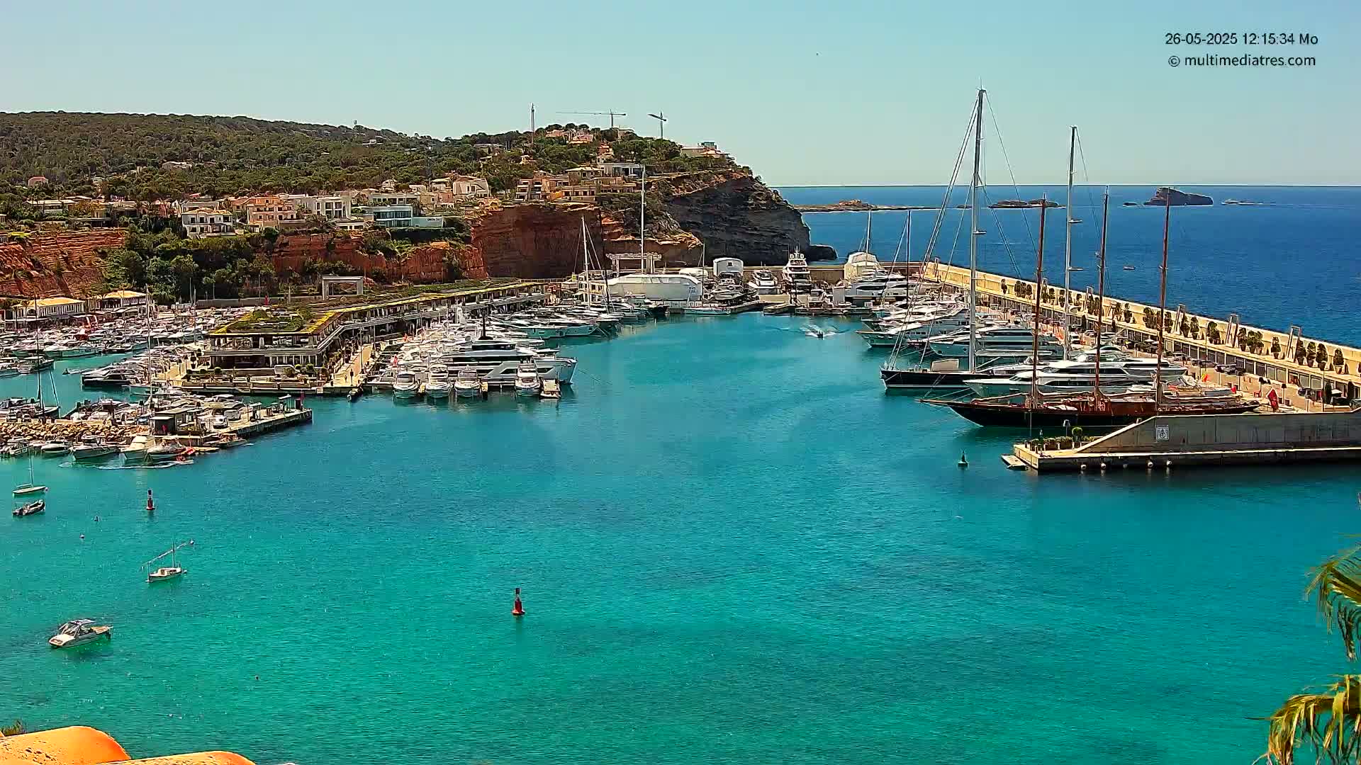 A marina filled with yachts and boats is nestled into a coastline on a sunny day with clear turquoise water.