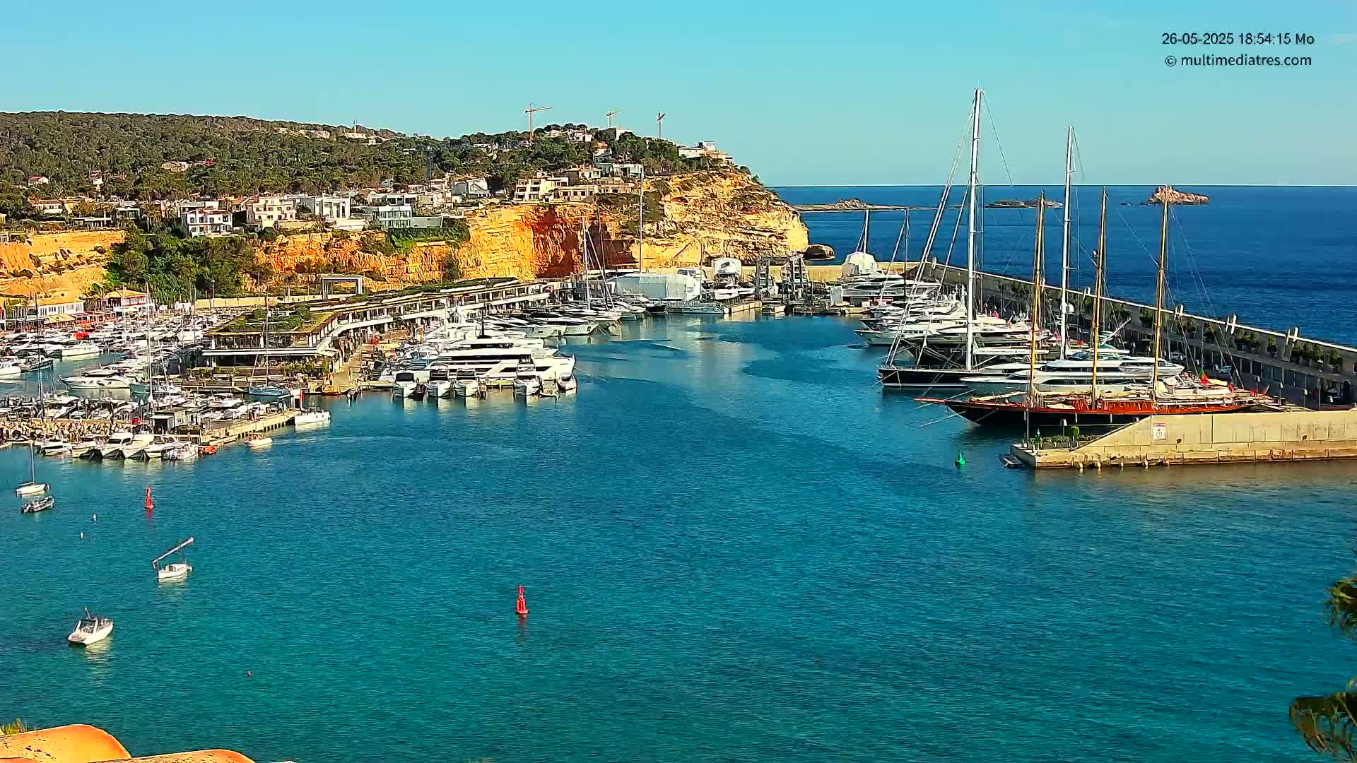 A marina filled with numerous yachts and sailboats is nestled against a sunny, hilly coastline.