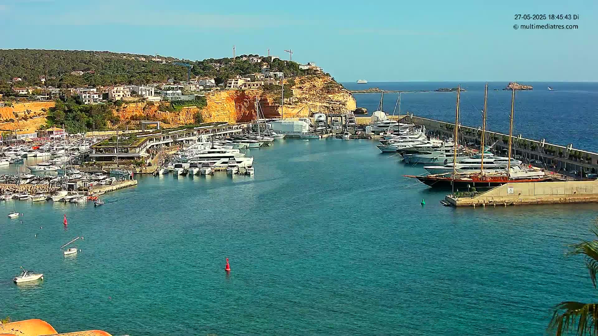 A marina filled with yachts and sailboats is nestled into a rocky coastline under a sunny sky.