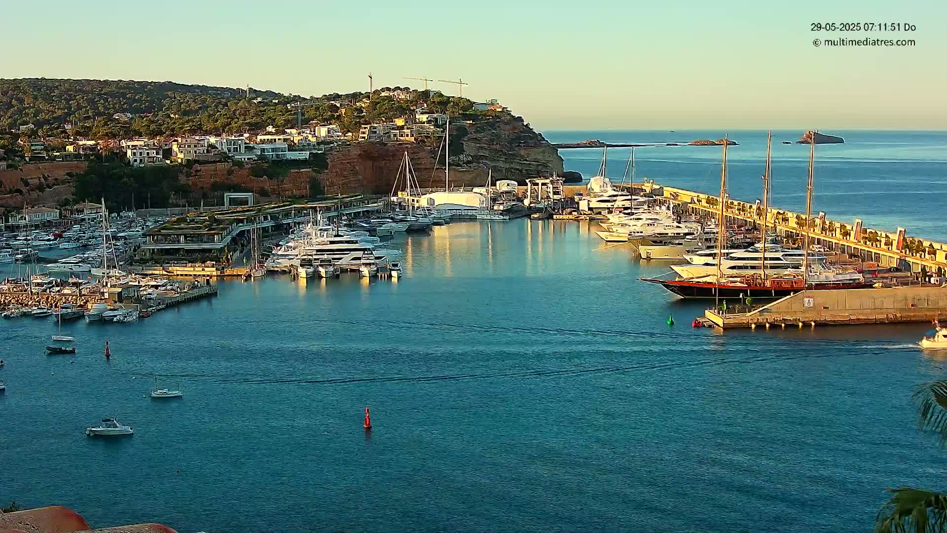 A calm harbor filled with numerous yachts and boats is nestled against a hillside of buildings under a clear, bright sky.