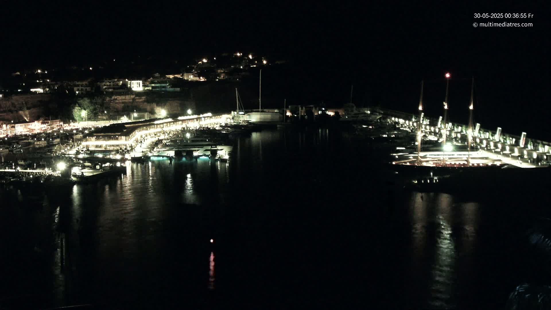 A nighttime view of a harbor filled with boats, lit buildings along the shoreline, and a dark, calm body of water reflecting the lights.