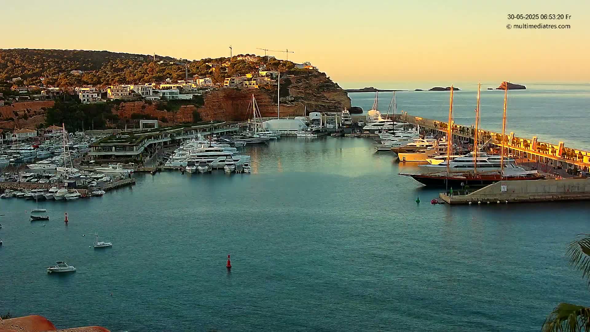 A calm harbor filled with numerous yachts and boats is nestled against a cliffside town at sunset.