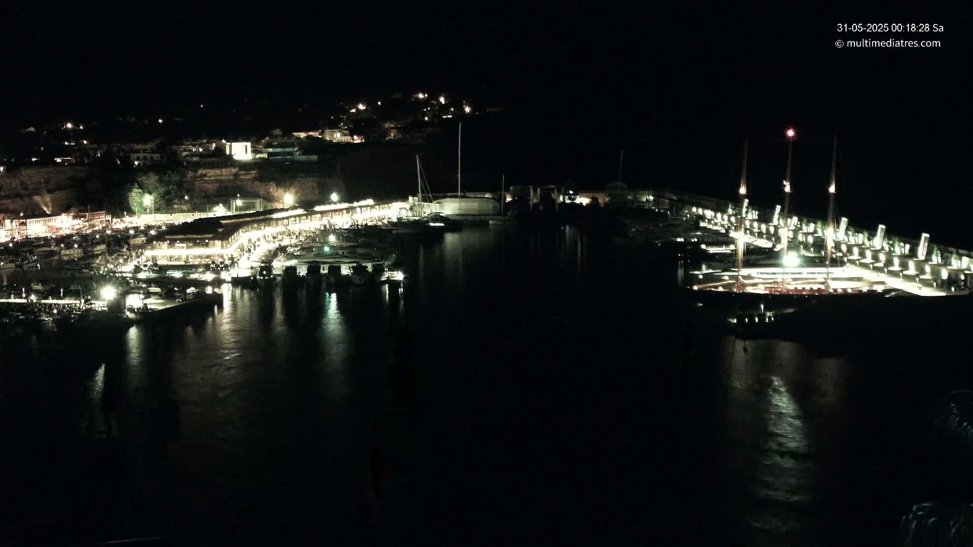 A nighttime view of a harbor filled with boats, illuminated buildings along the shoreline, and a dark, calm body of water.