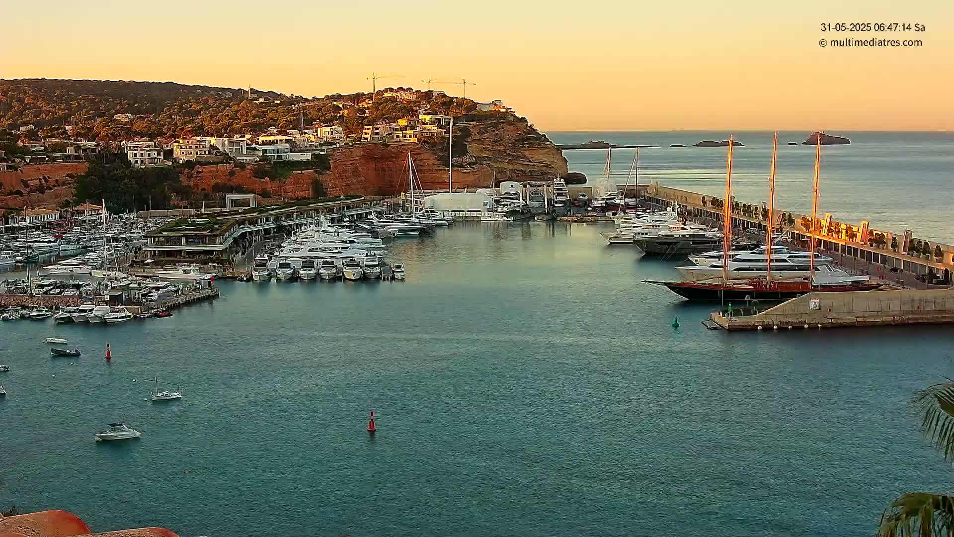 A calm harbor filled with numerous yachts and boats at sunset, nestled against a cliffside town.