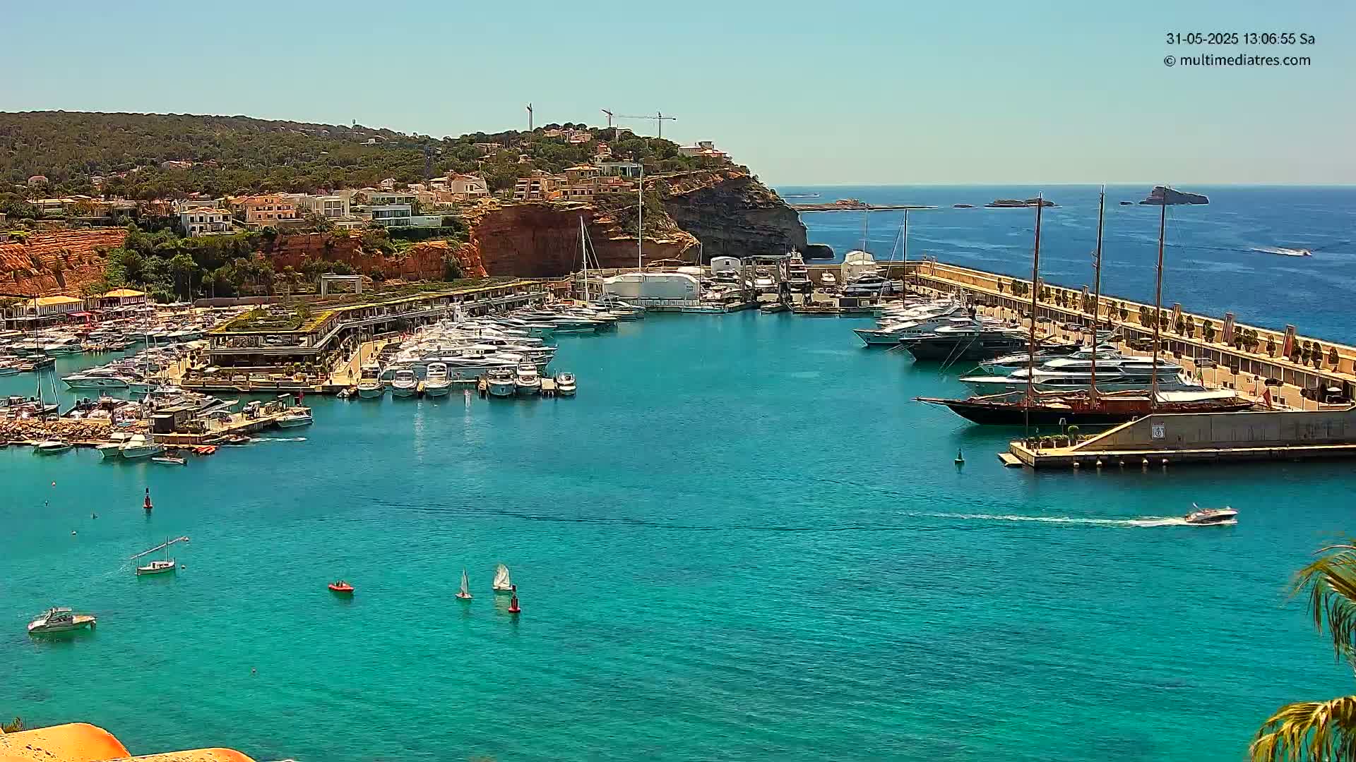 A marina filled with yachts and sailboats is nestled in a calm, turquoise bay on a sunny day, with a hillside of houses and vegetation rising behind it.