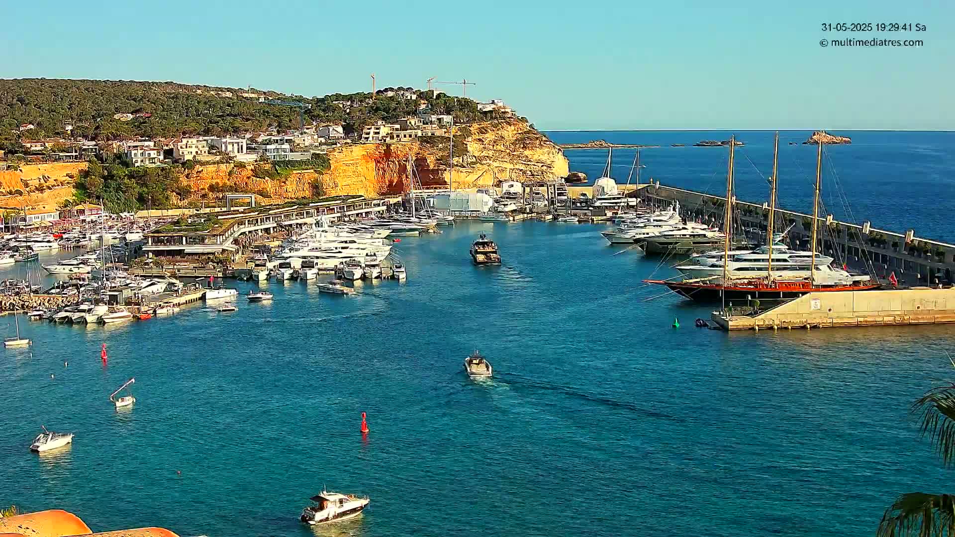 A marina filled with numerous yachts and boats is situated against a backdrop of a sunny, cliffside town.