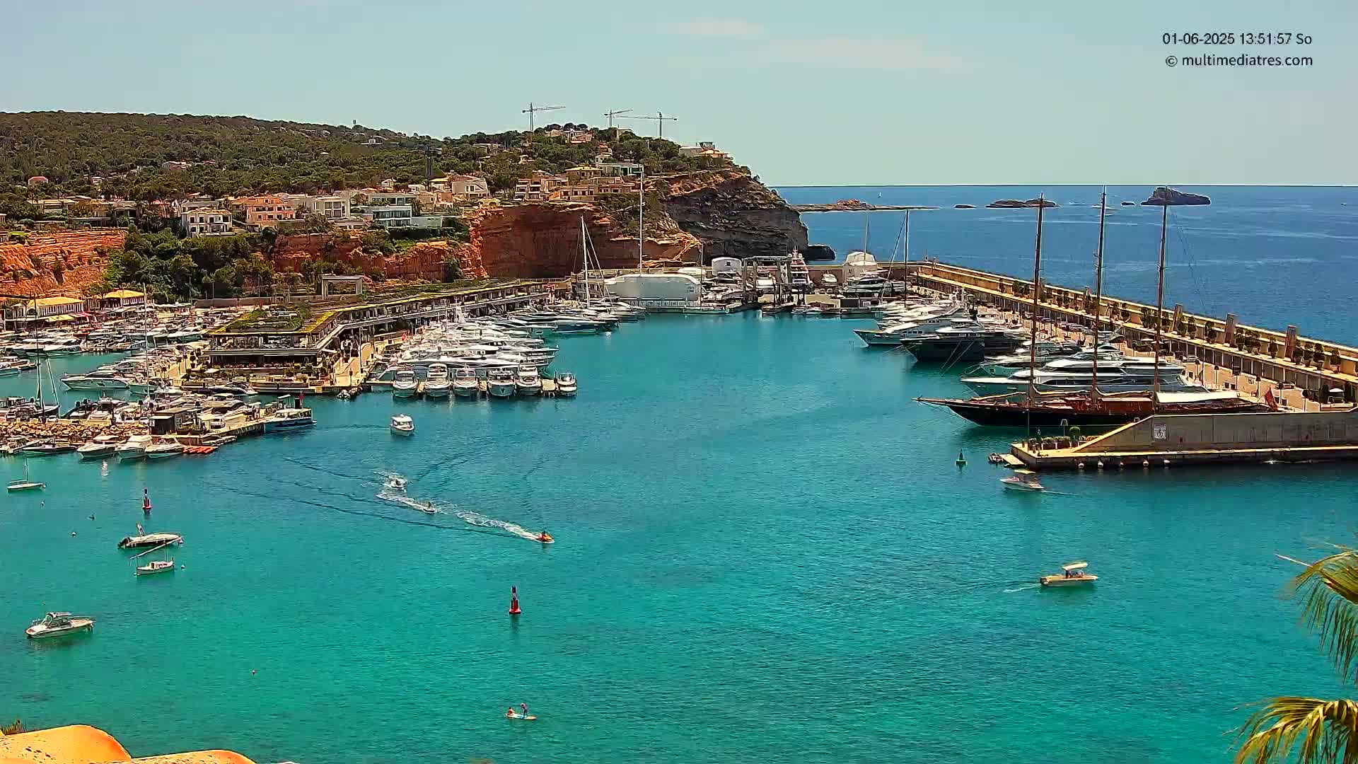 A marina filled with numerous yachts and boats is nestled against a red-cliffed coastline under a clear, sunny sky.