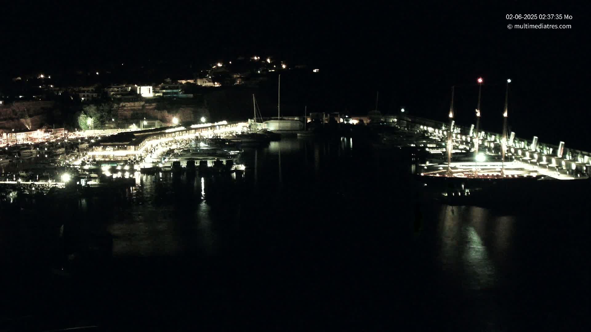 A nighttime view of a harbor filled with boats, lit buildings along the shore, and a hill in the background under a clear, dark sky.