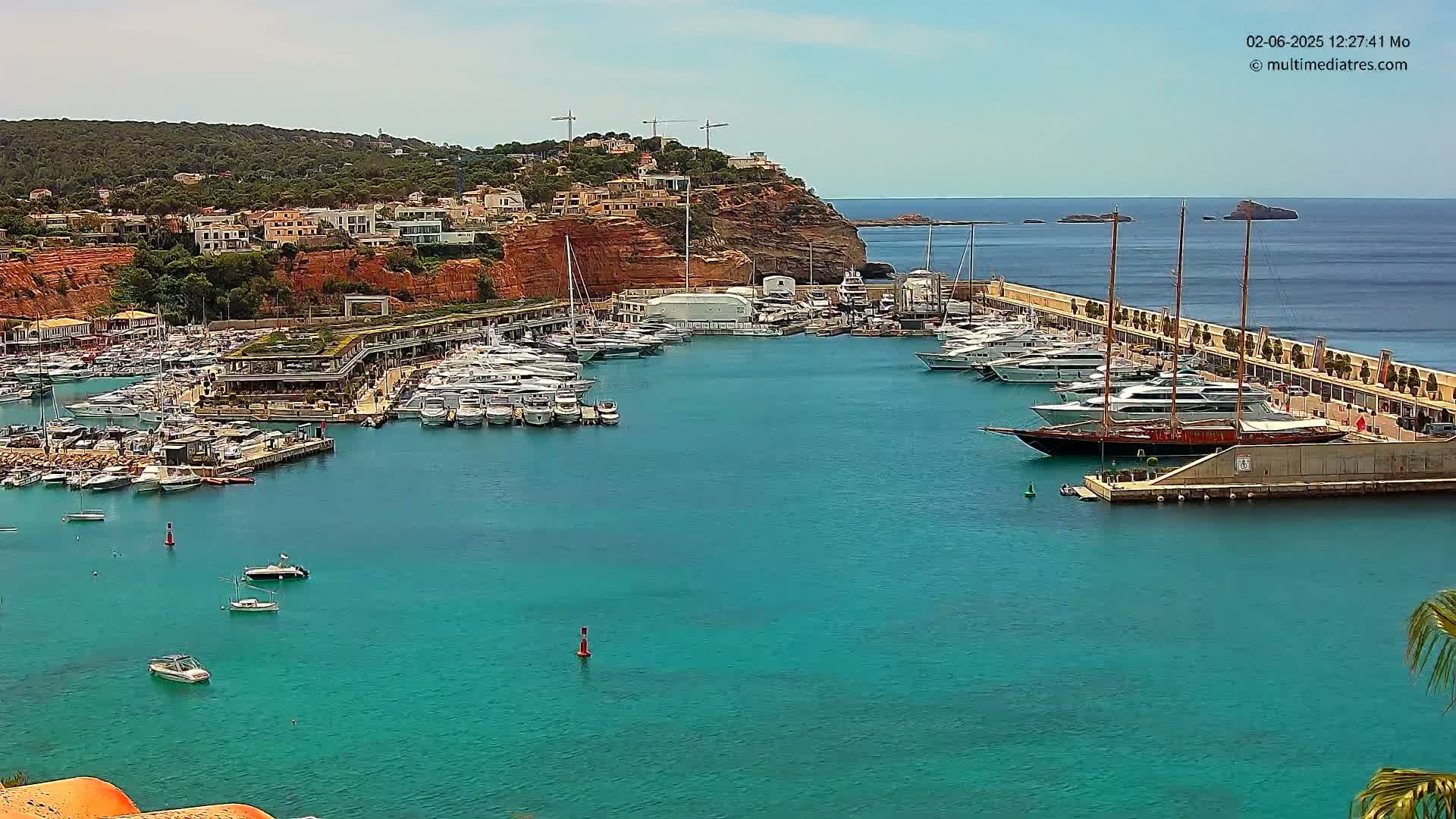 A marina filled with numerous yachts and boats is nestled into a calm, turquoise bay, backed by a hillside of buildings and red cliffs under a clear sky.