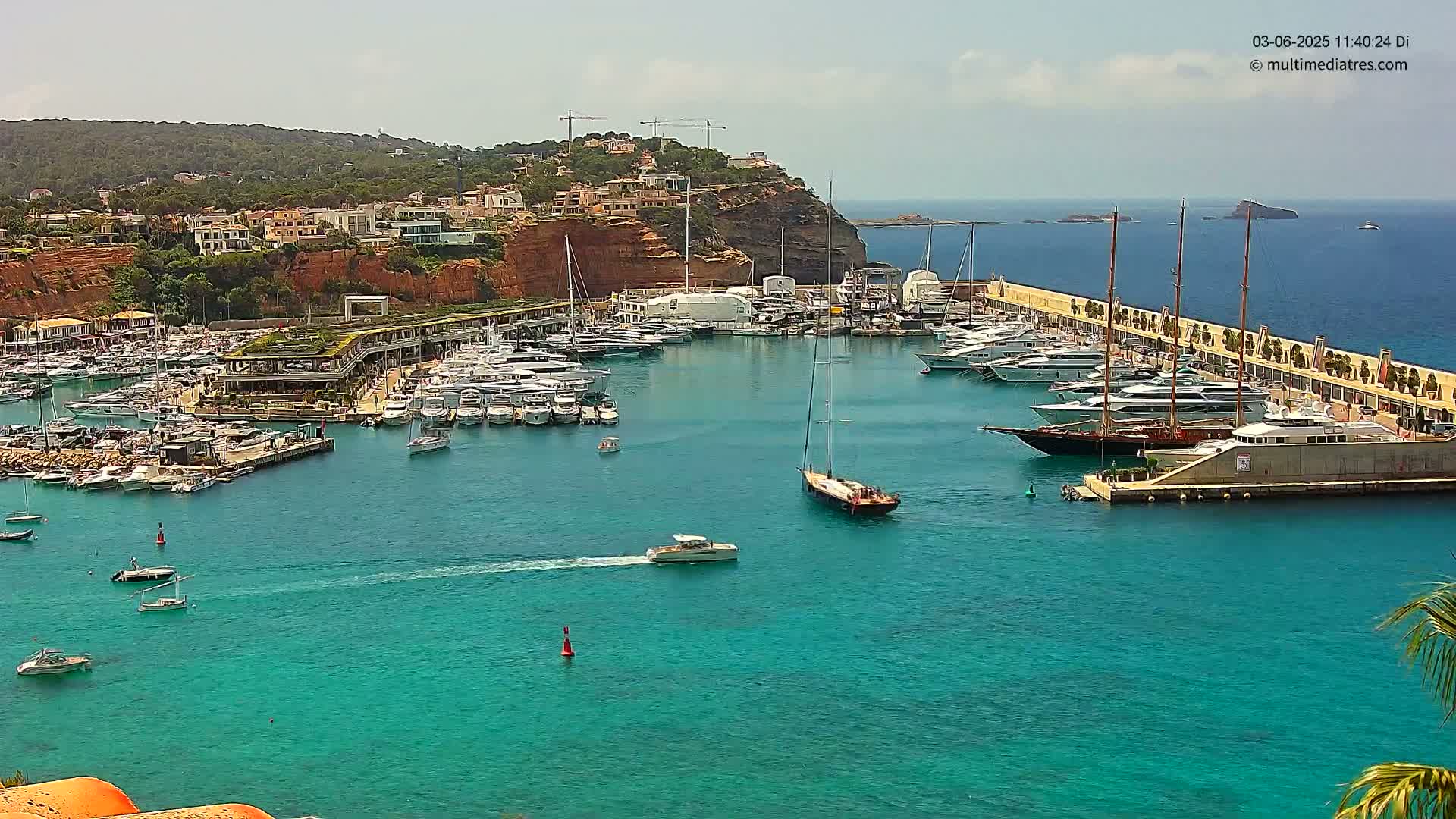 A marina filled with yachts and boats is situated in a calm, turquoise harbor next to a red cliffside town under a partly sunny sky.