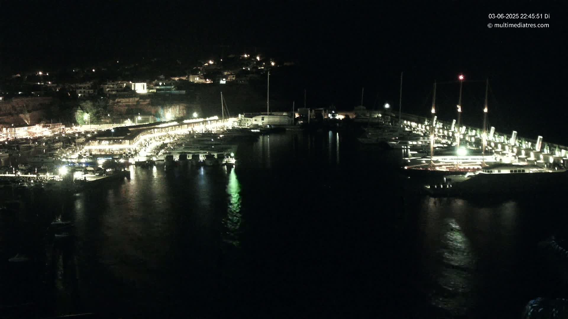 A brightly lit marina filled with boats at night, situated below a dark hillside with buildings.