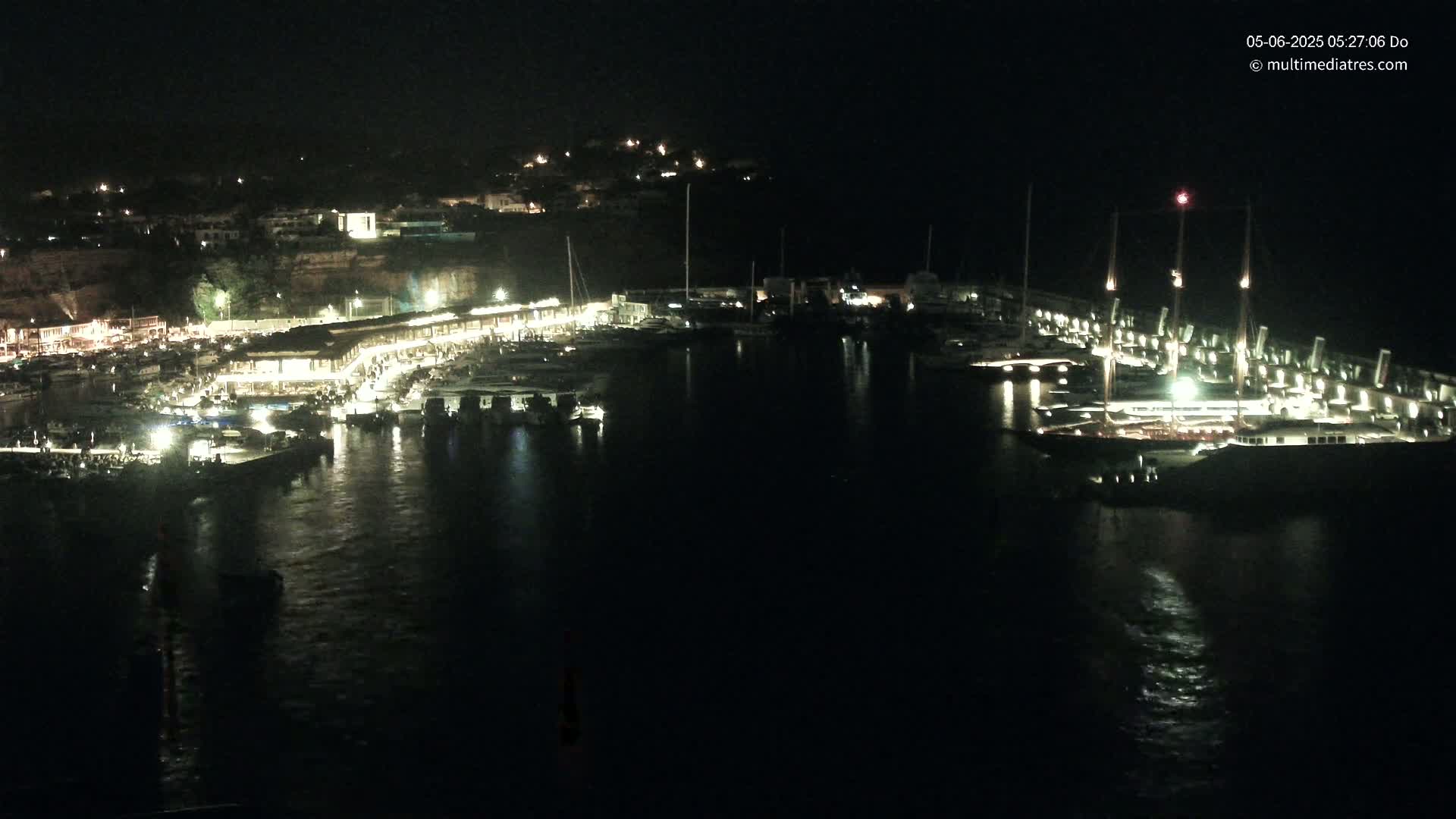 A nighttime view of a harbor filled with boats, lit up by building lights and docked vessel lights, against a dark, hilly background.