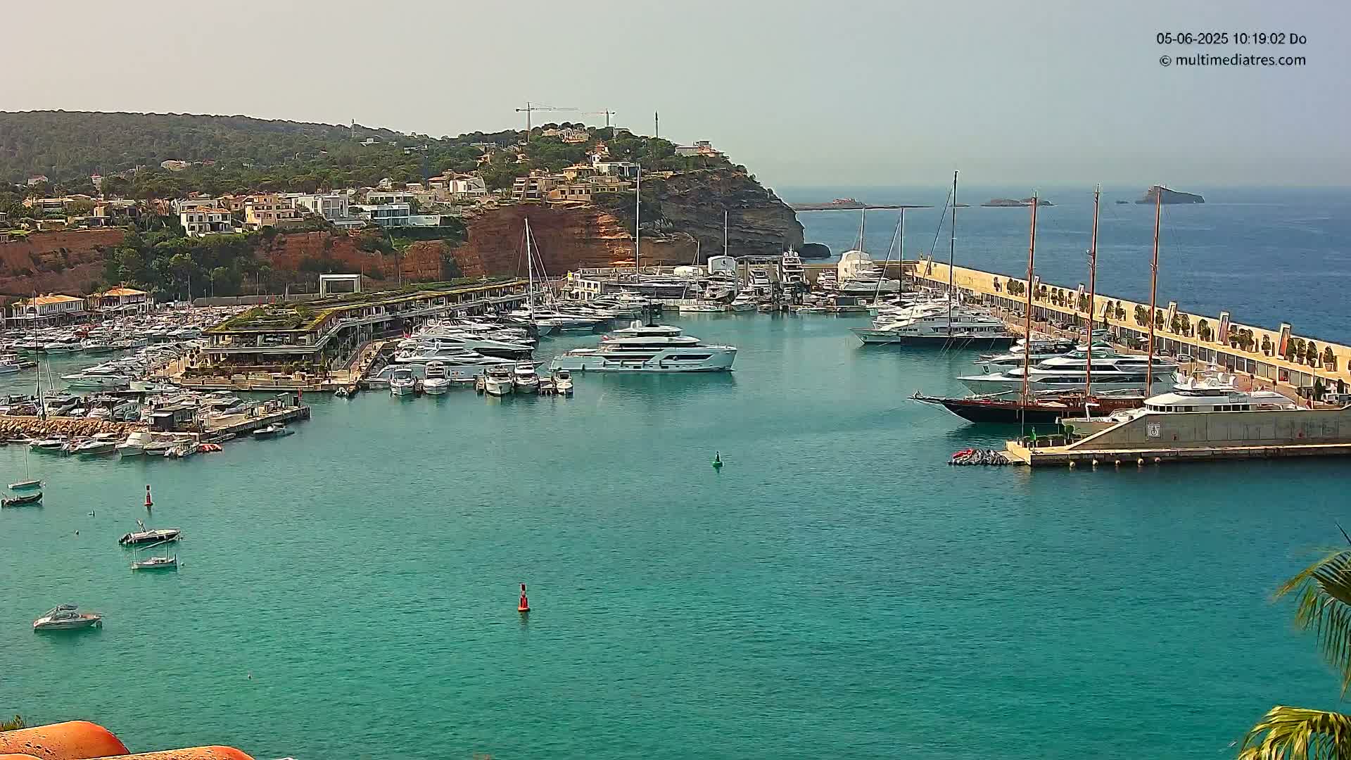 A marina filled with yachts and boats is nestled in a calm, turquoise bay next to a hillside town under a clear, sunny sky.