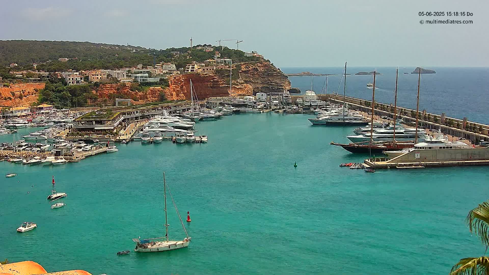 A marina filled with numerous boats and yachts is situated next to a red-cliffed coast under a mostly clear sky.