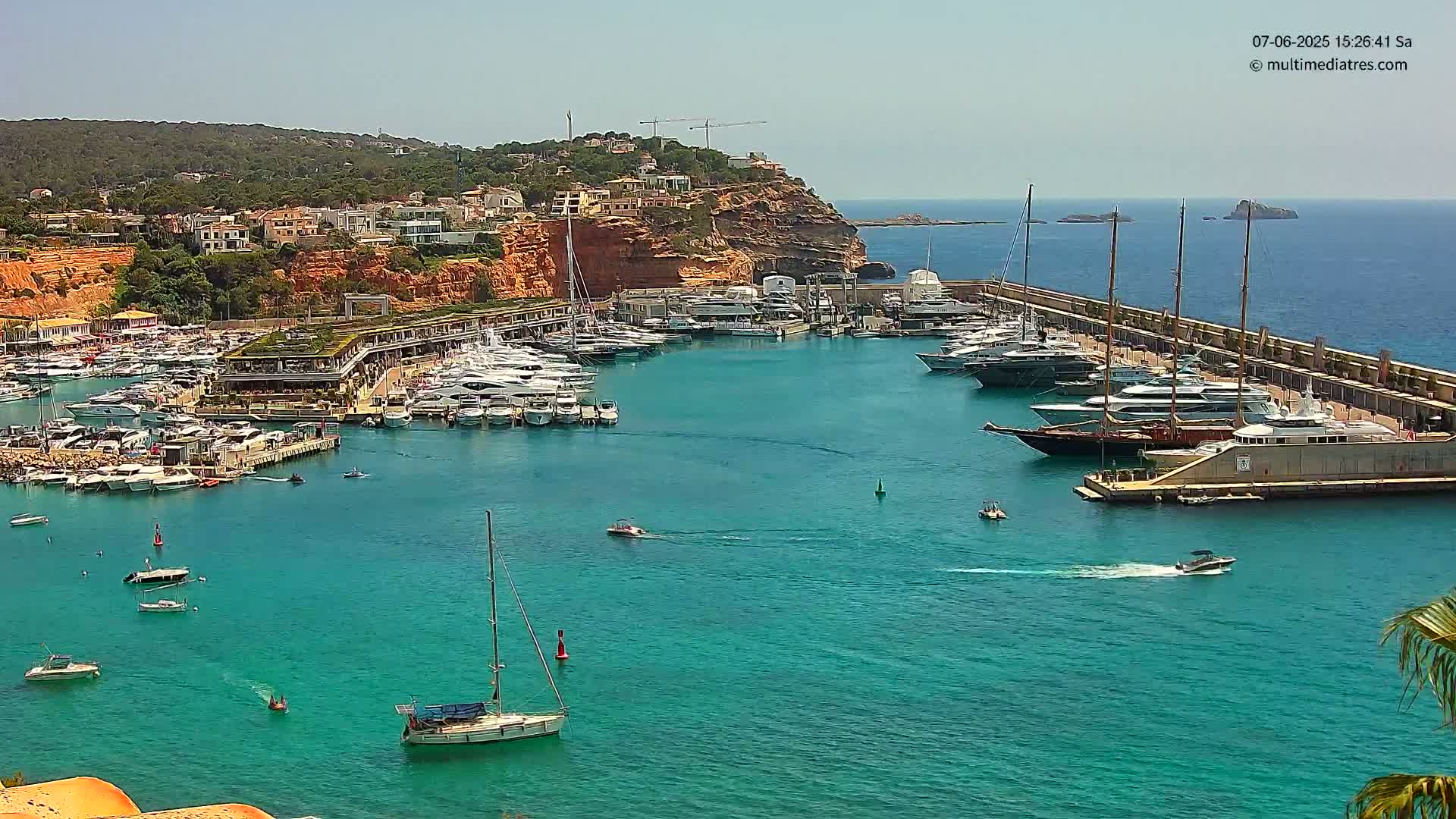 A marina filled with numerous yachts and boats is nestled in a calm, turquoise bay on a sunny day, with a hillside of buildings and red cliffs visible in the background.