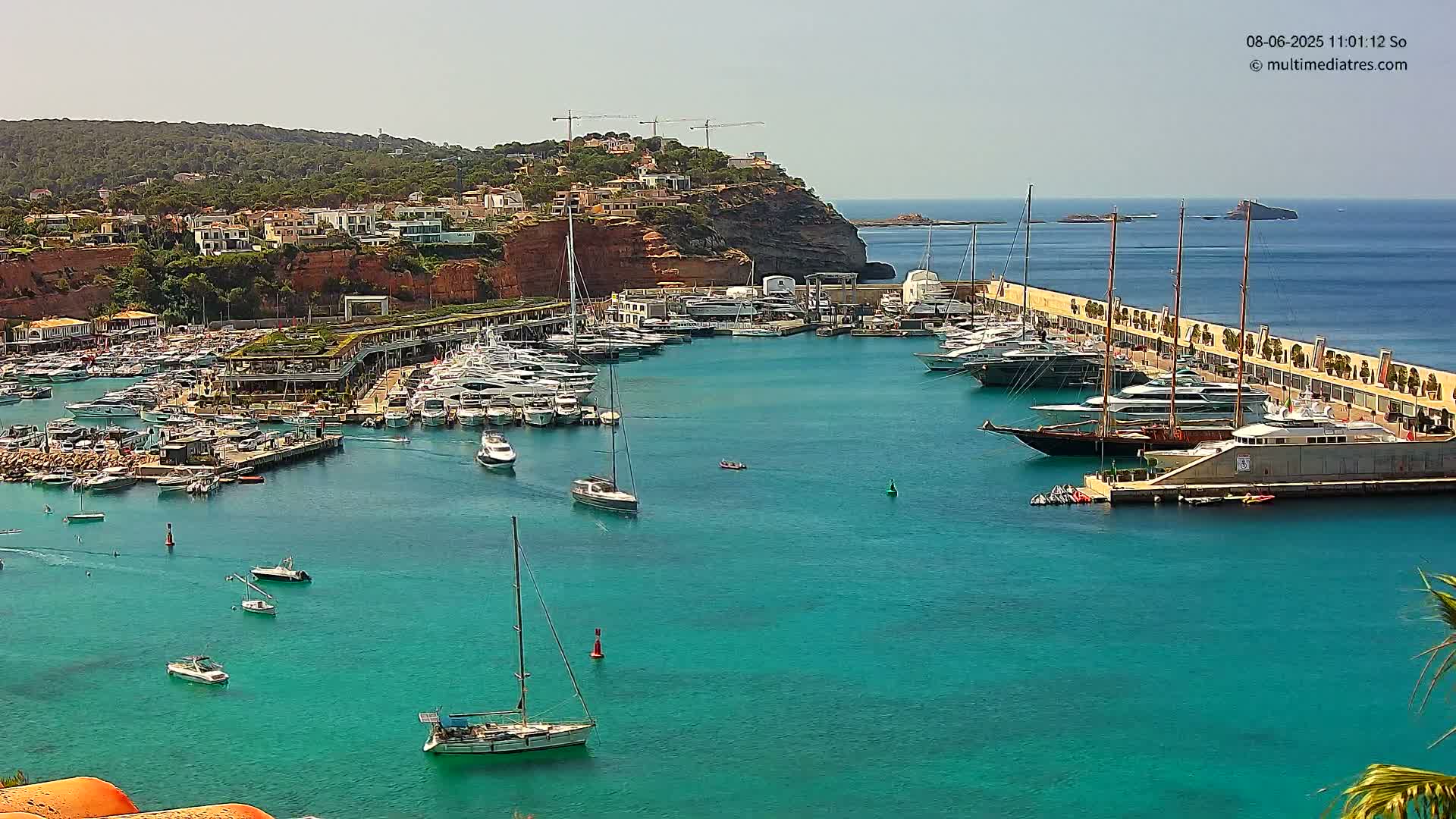 A marina filled with numerous yachts and sailboats is nestled in a turquoise bay, under a clear, sunny sky, with a hillside of buildings and vegetation in the background.
