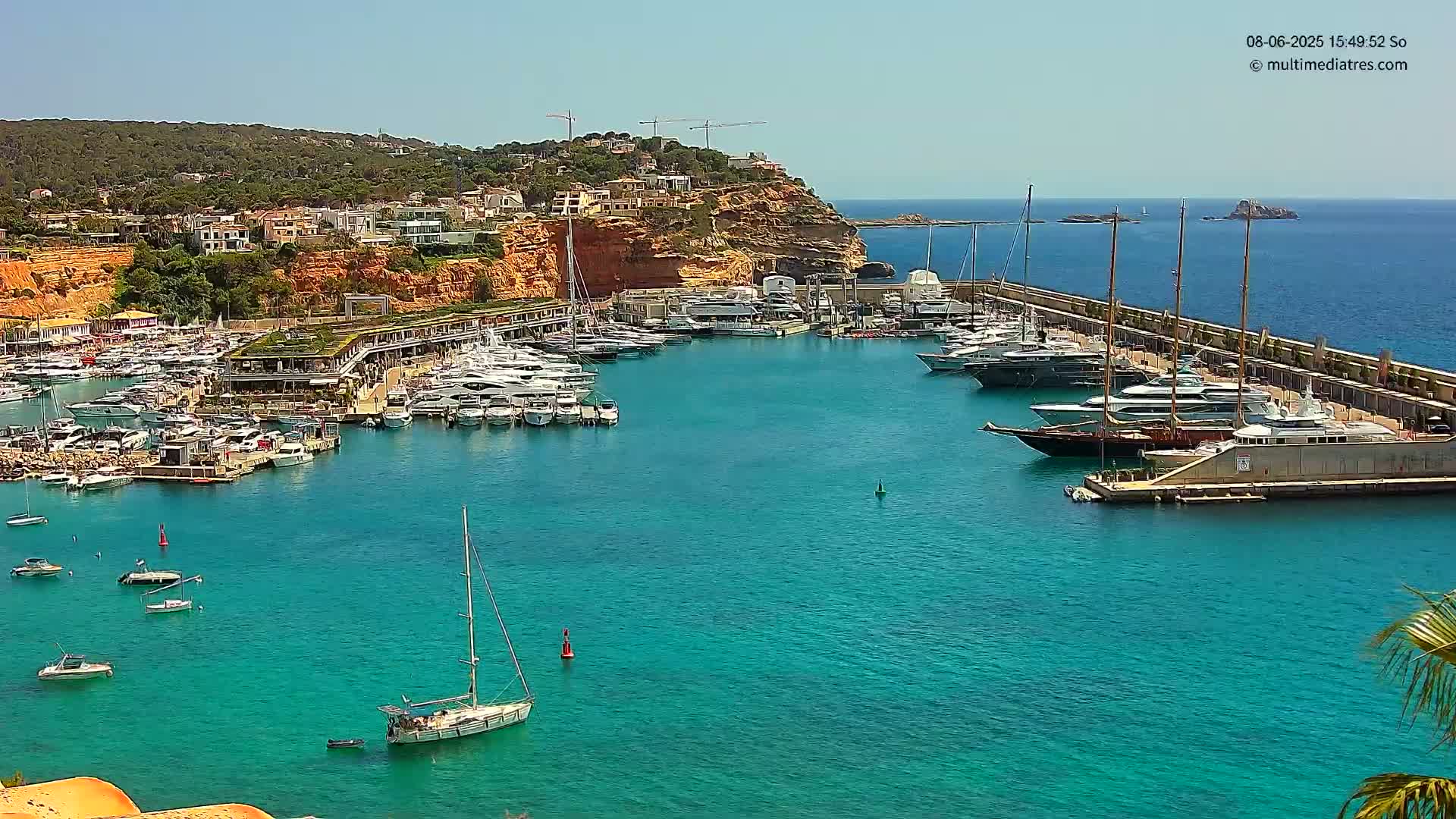 A harbor filled with numerous yachts and sailboats is nestled along a rocky coastline under a clear, sunny sky.