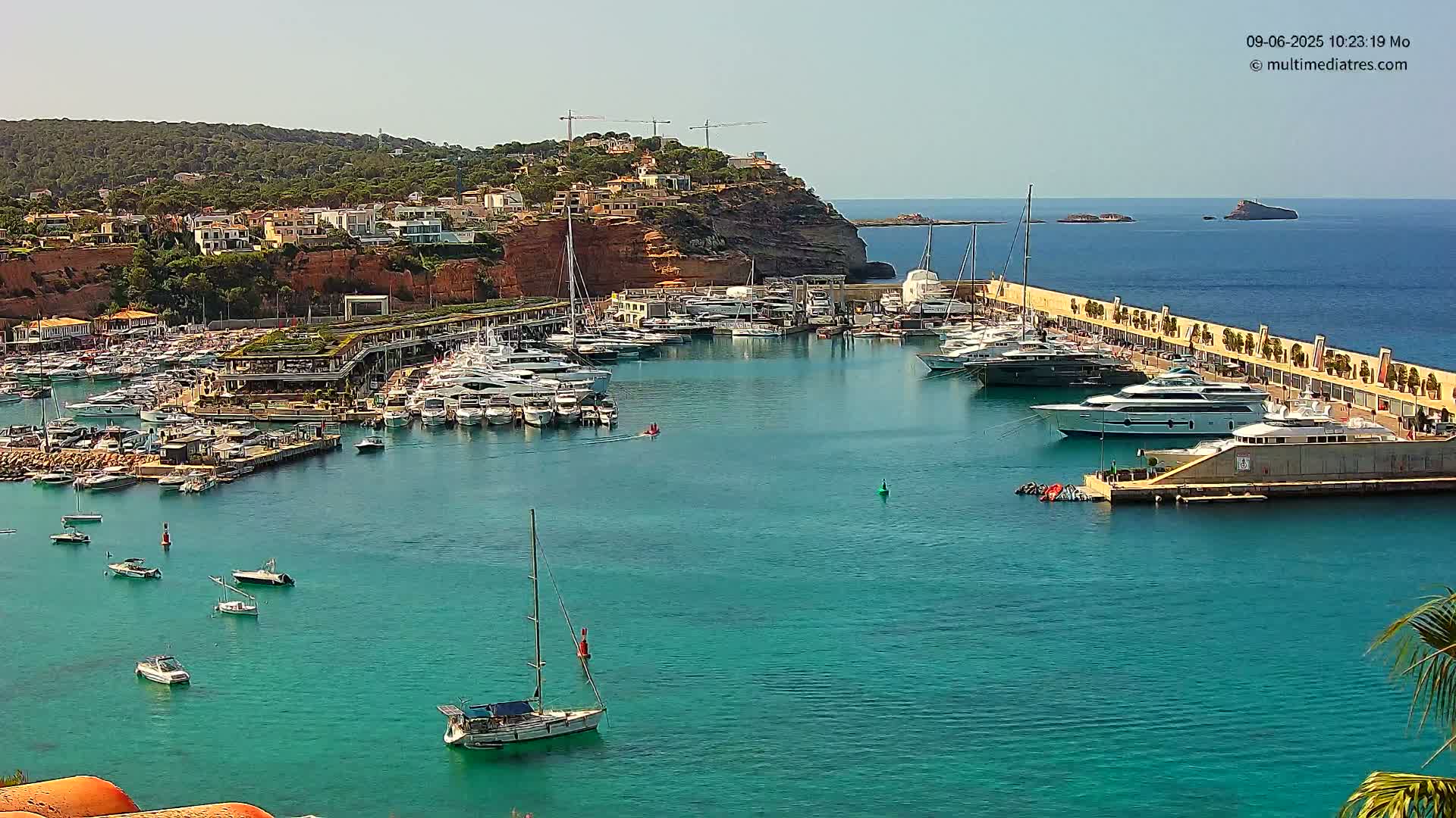 A marina filled with numerous yachts and boats is nestled in a turquoise bay under a clear sunny sky, backed by a hillside of buildings and lush greenery.