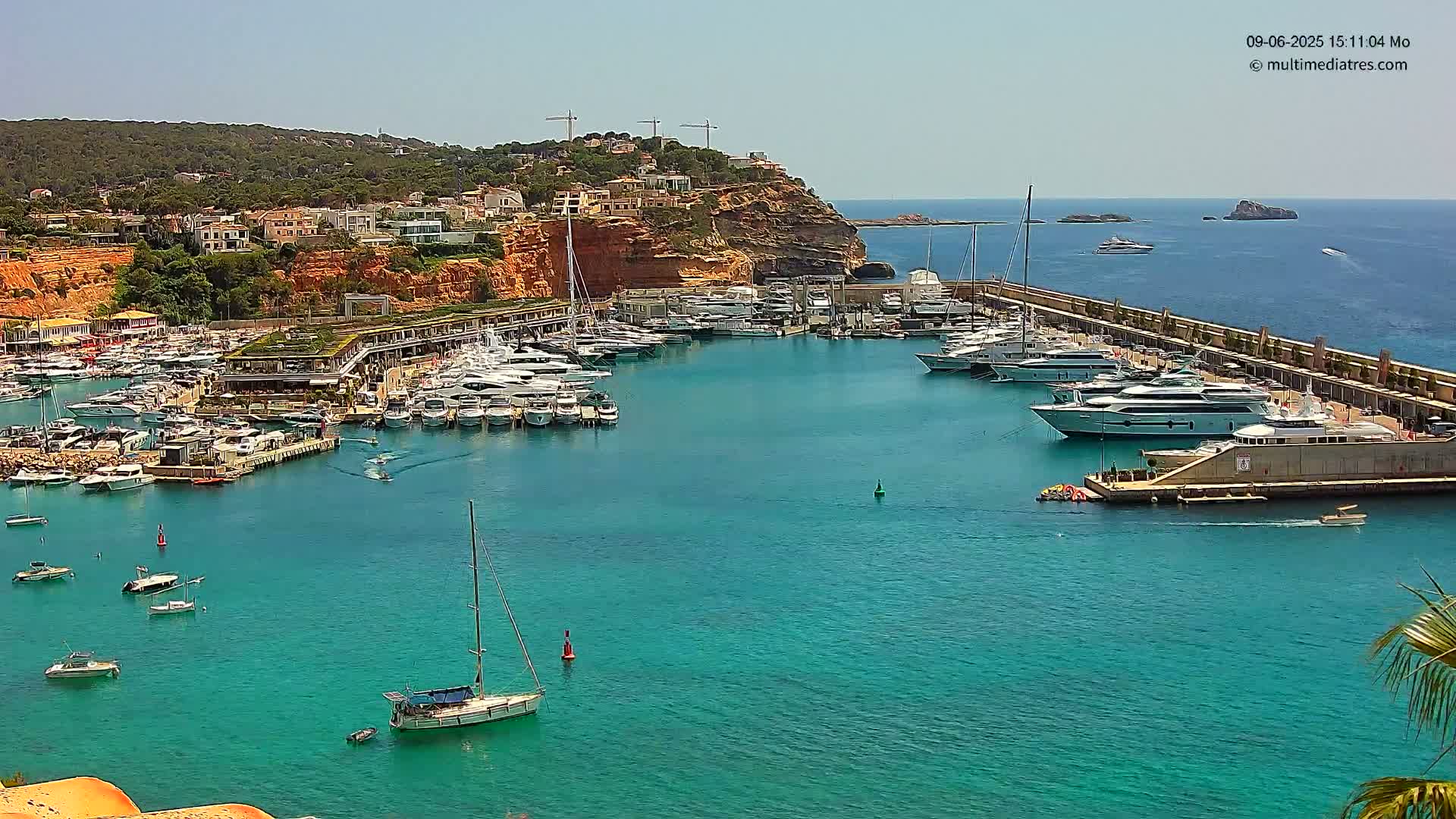 A marina filled with yachts and sailboats is nestled in a turquoise bay on a sunny day, with a hillside of buildings visible in the background.