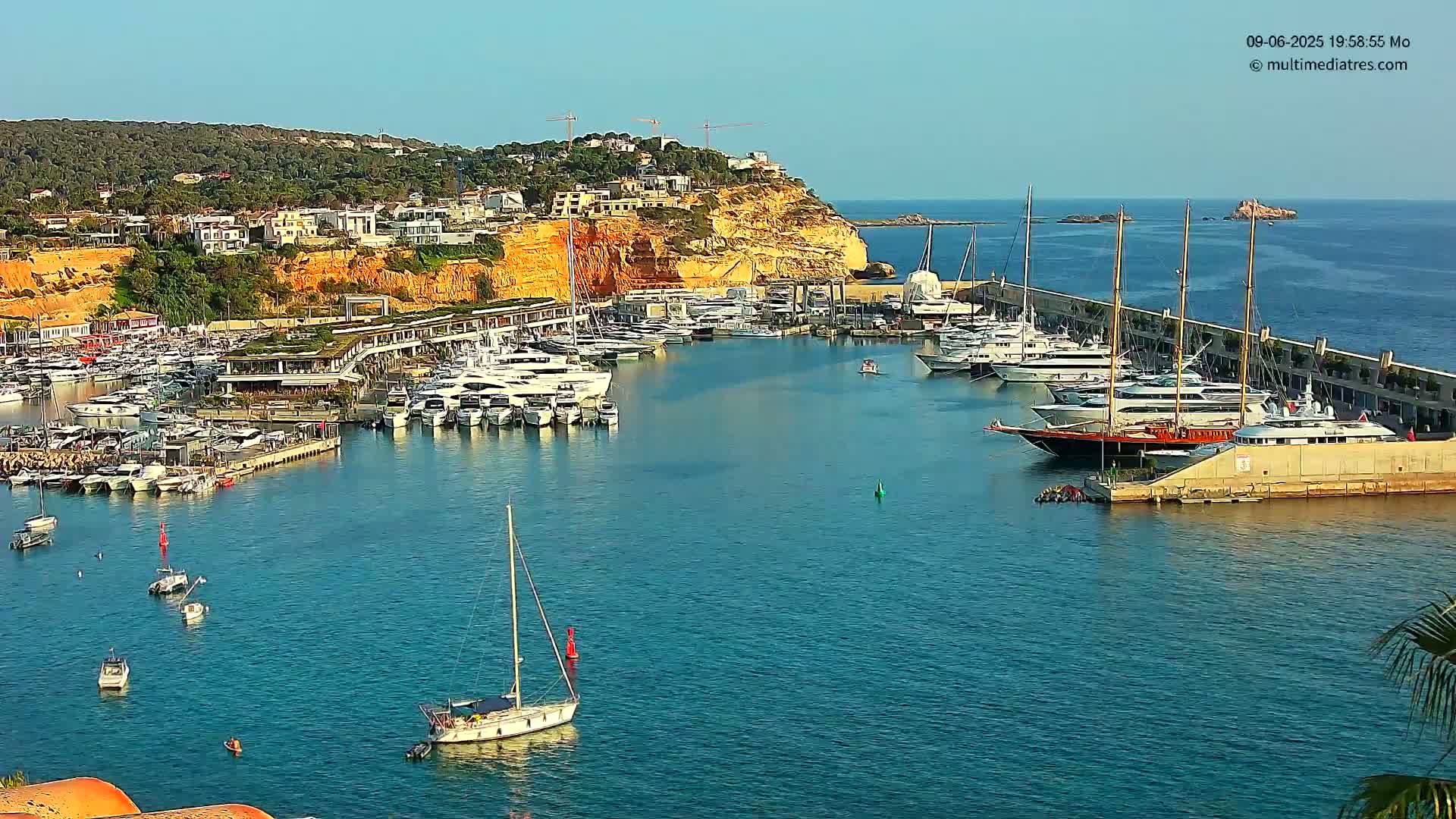 A harbor filled with yachts and sailboats is nestled against a sunny, hilly coastline.