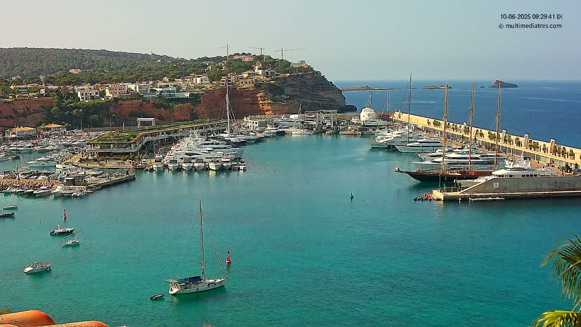 A marina filled with numerous yachts and sailboats is situated in a calm, clear turquoise bay against a backdrop of a hilly coastline under a sunny sky.