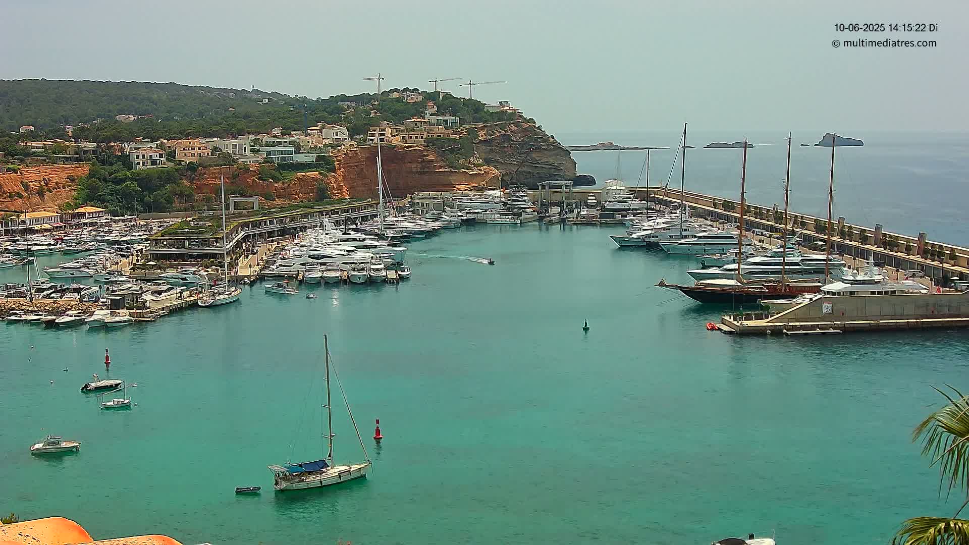 A marina filled with numerous yachts and sailboats is nestled into a calm, turquoise bay, bordered by a hillside of buildings under a clear, sunny sky.