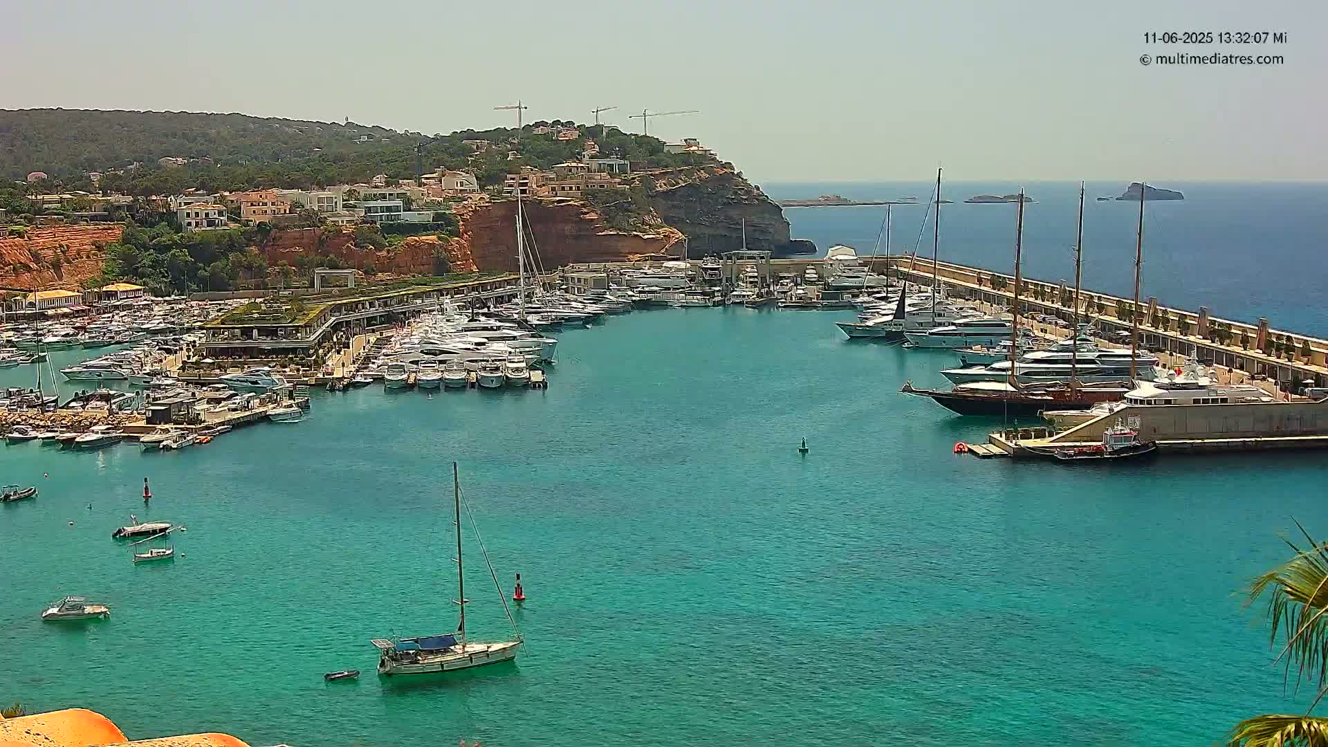 A marina filled with numerous yachts and boats is nestled in a turquoise bay, backed by a hillside of buildings and under a clear, sunny sky.