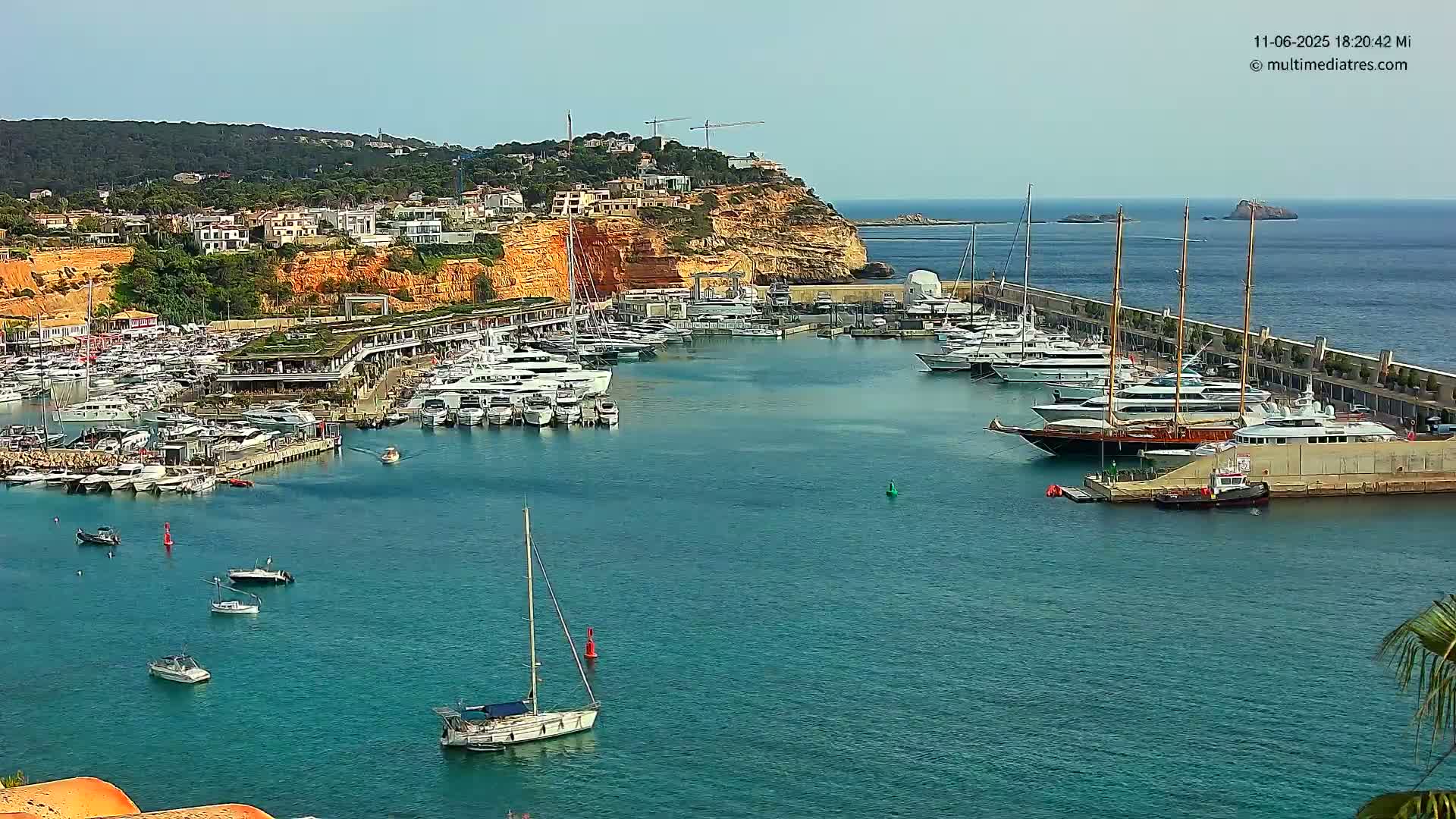 A harbor filled with numerous yachts and sailboats is situated against a backdrop of a sunny, coastal town nestled on a cliffside.