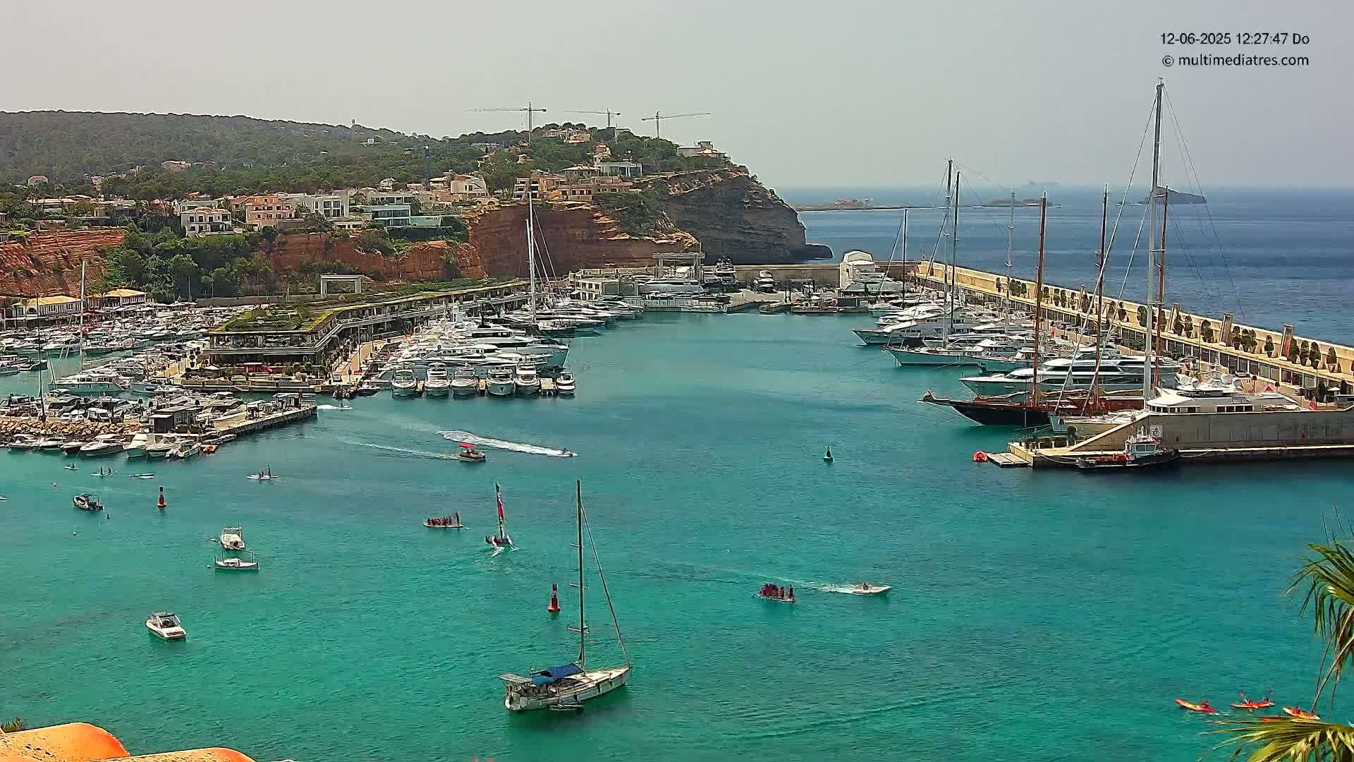A marina filled with numerous yachts and sailboats is situated on a calm, turquoise body of water next to a red-cliff hillside on a sunny day.